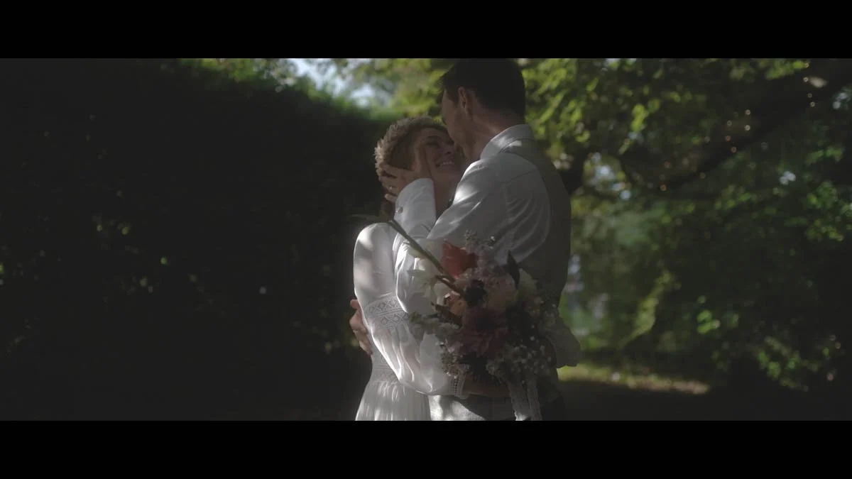 A couple share an intimate embrace beneath softly dappled woodland light as the bride smiles up at her partner while holding a bouquet of muted pinks greens and delicate florals that blend gently into the dreamy cinematic shadows around them.