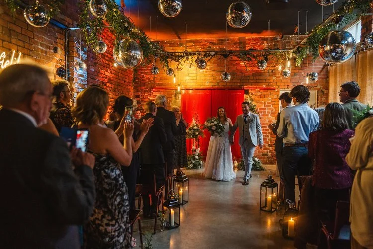 A stylish industrial wedding ceremony at The Village Green Events, Sheffield. The newlyweds make their grand entrance as guests applaud beneath a canopy of disco balls, twinkling lanterns, and lush greenery. The warm lighting, exposed brick walls, an