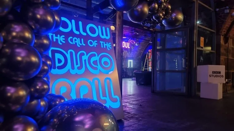 My Colour is Gold illuminated sign reading 'Follow the call of the disco ball' in glowing blue, styled with metallic balloons for a fun party atmosphere.