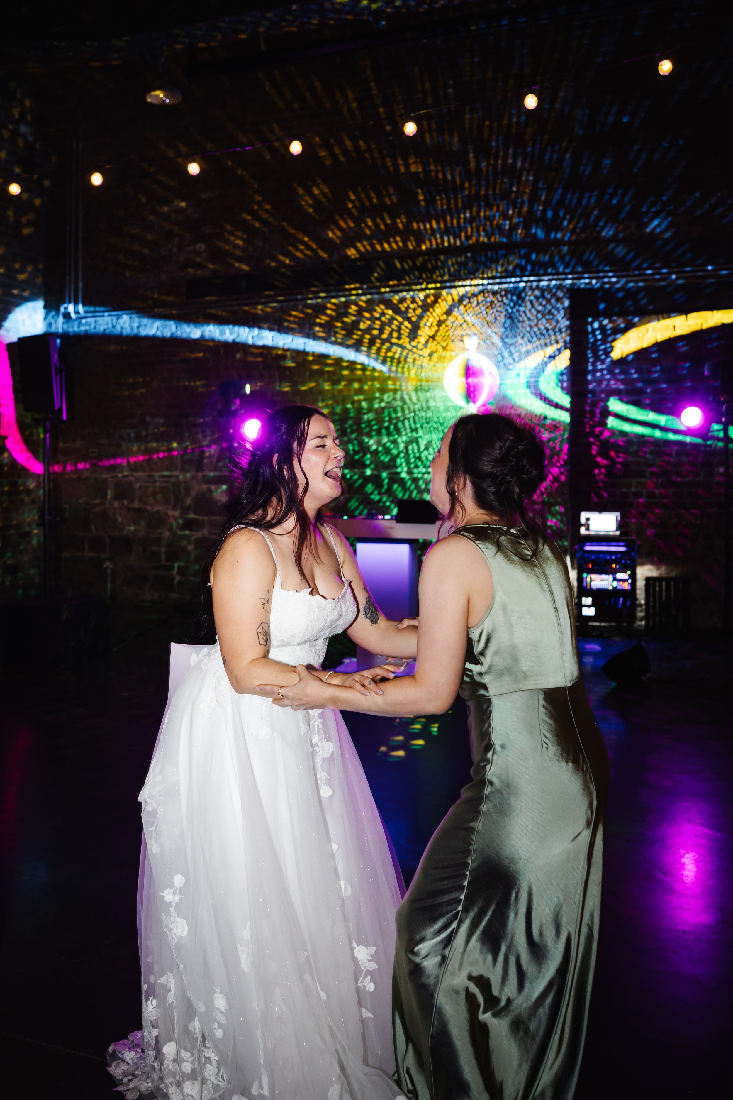 Marie and one of her bridesmaids holding each others arms and signing in front of some colourful disco lights on the dance floor.