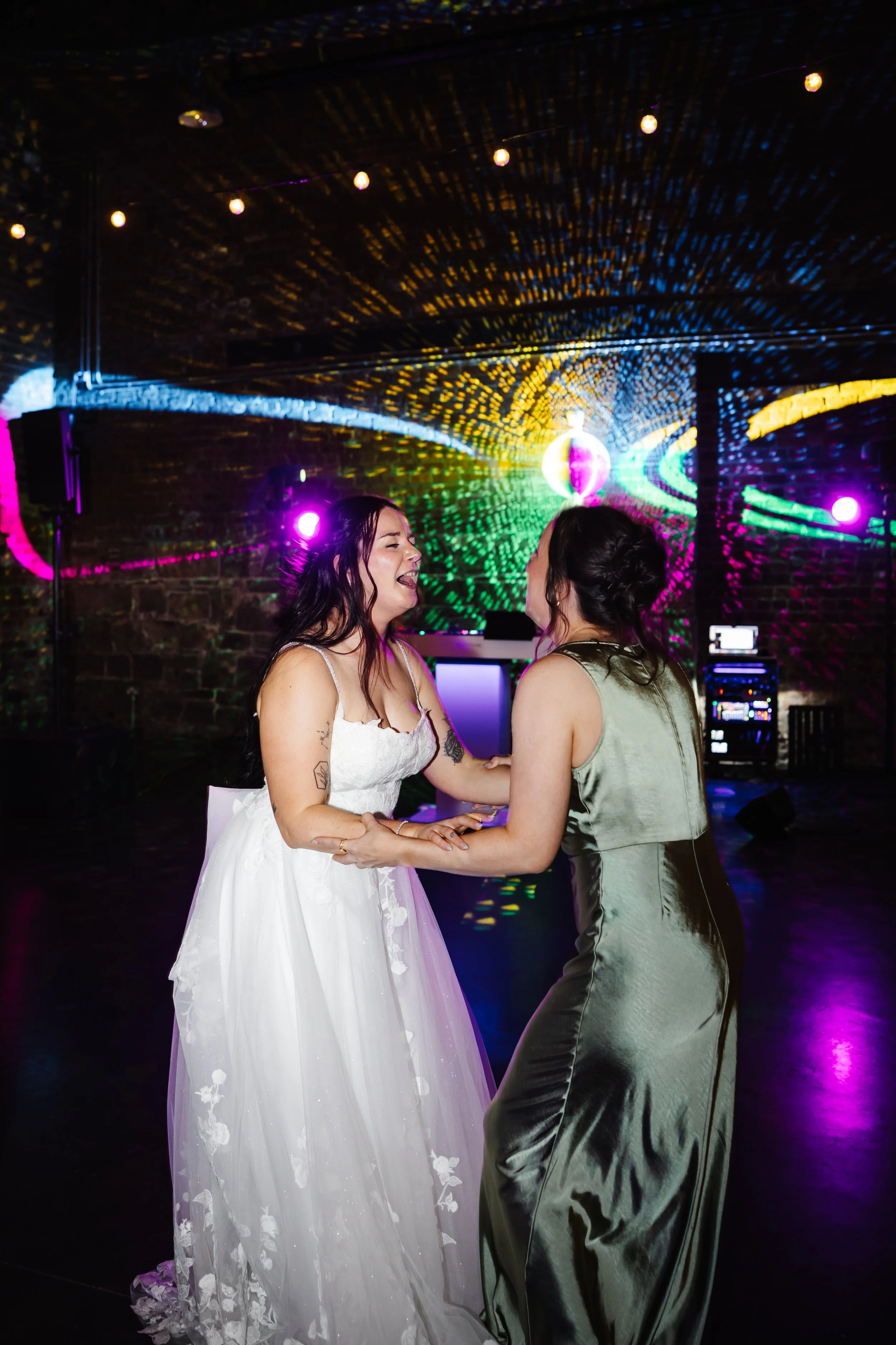 Marie and one of her bridesmaids holding each others arms and signing in front of some colourful disco lights on the dance floor.