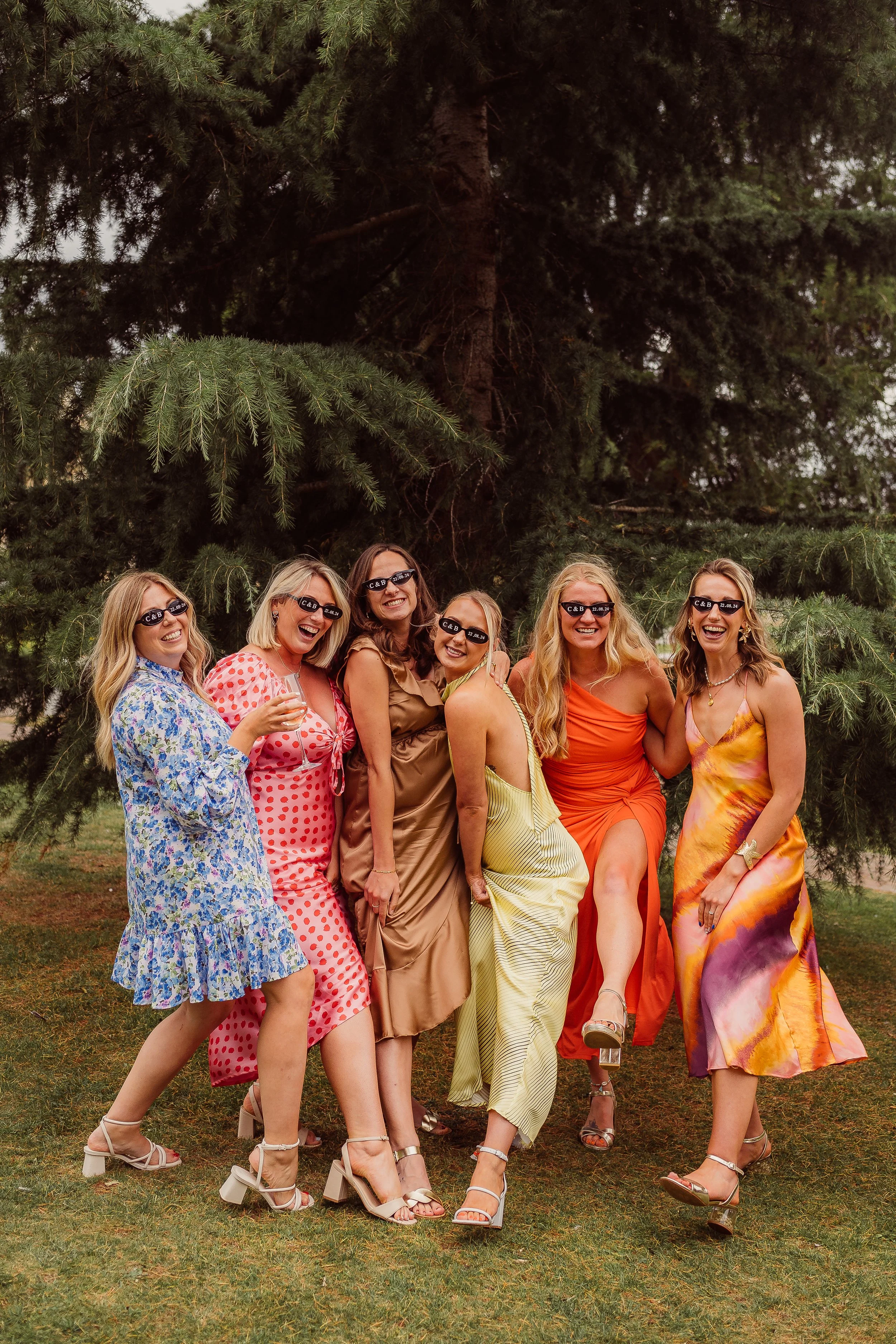 A group of friends in bold, colourful dresses celebrates together under a towering evergreen tree. They stand close in a playful pose with their legs kicked forward and matching sunglasses that add a fun, coordinated touch. The mood is joyful and rel