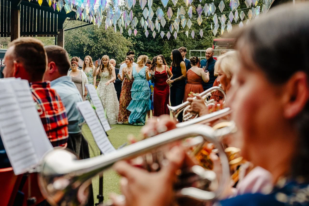 Guests stand gathered under shimmering iridescent decorations as a lively brass band plays in the foreground with trumpets and sheet music filling the frame while colourful dressed friends smile and sway creating a vibrant open air celebration full o