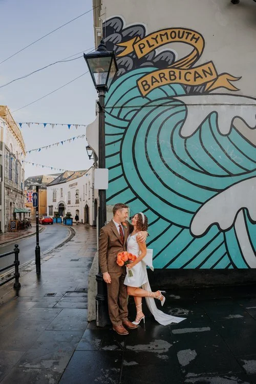 A newly married couple pose together beside a vibrant coastal mural in Plymouth Barbican, captured by Joshua Humphrey Photography. The bride lifts one foot playfully while holding a bright bouquet and the groom smiles at her, creating a fun relaxed w
