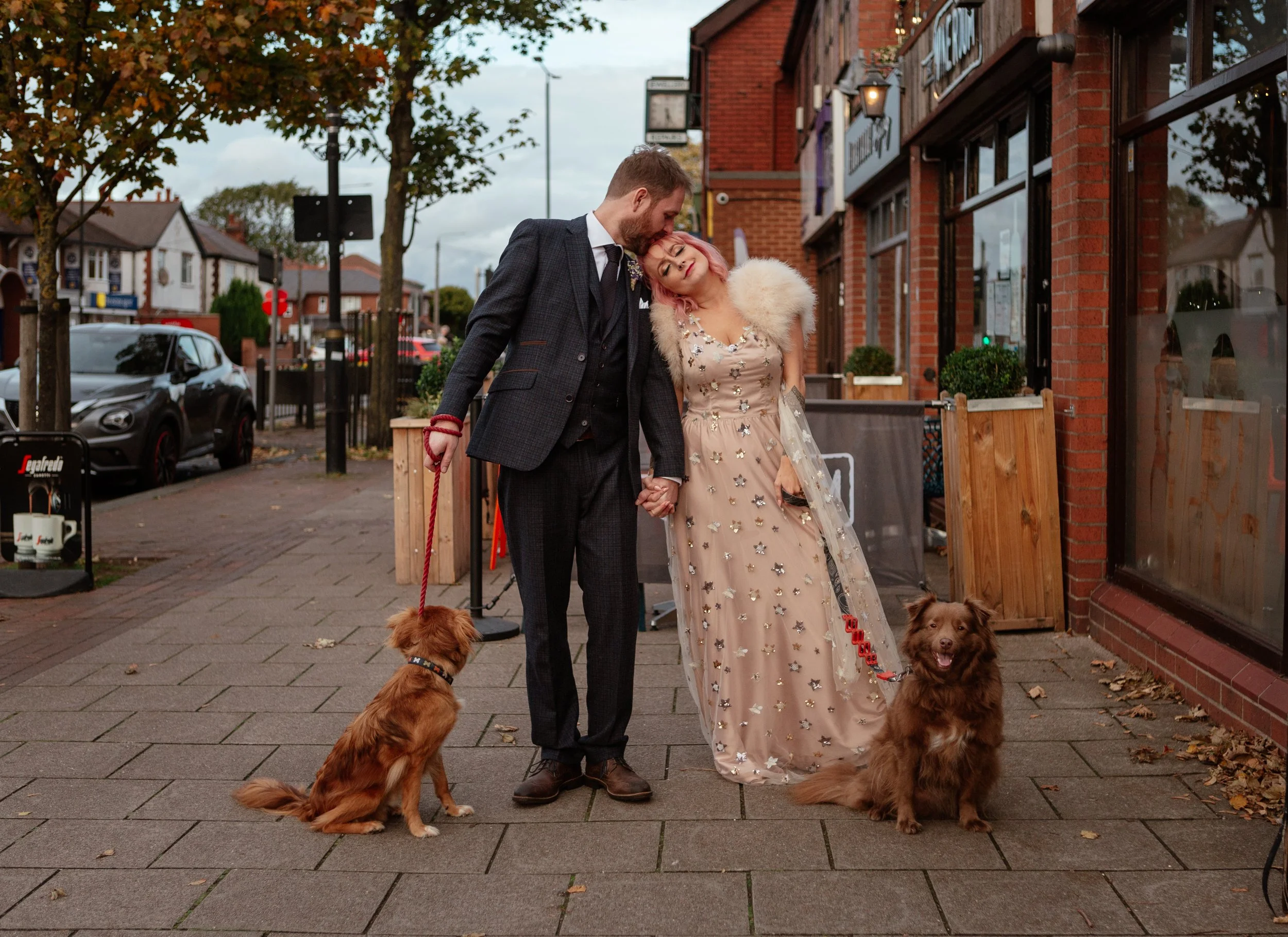 A charming, personality-filled wedding portrait captured by Laura Wilson Photography, showing a couple standing hand-in-hand on a suburban high street with their two dogs. The groom, dressed in a textured dark suit, leans in to kiss the bride’s foreh