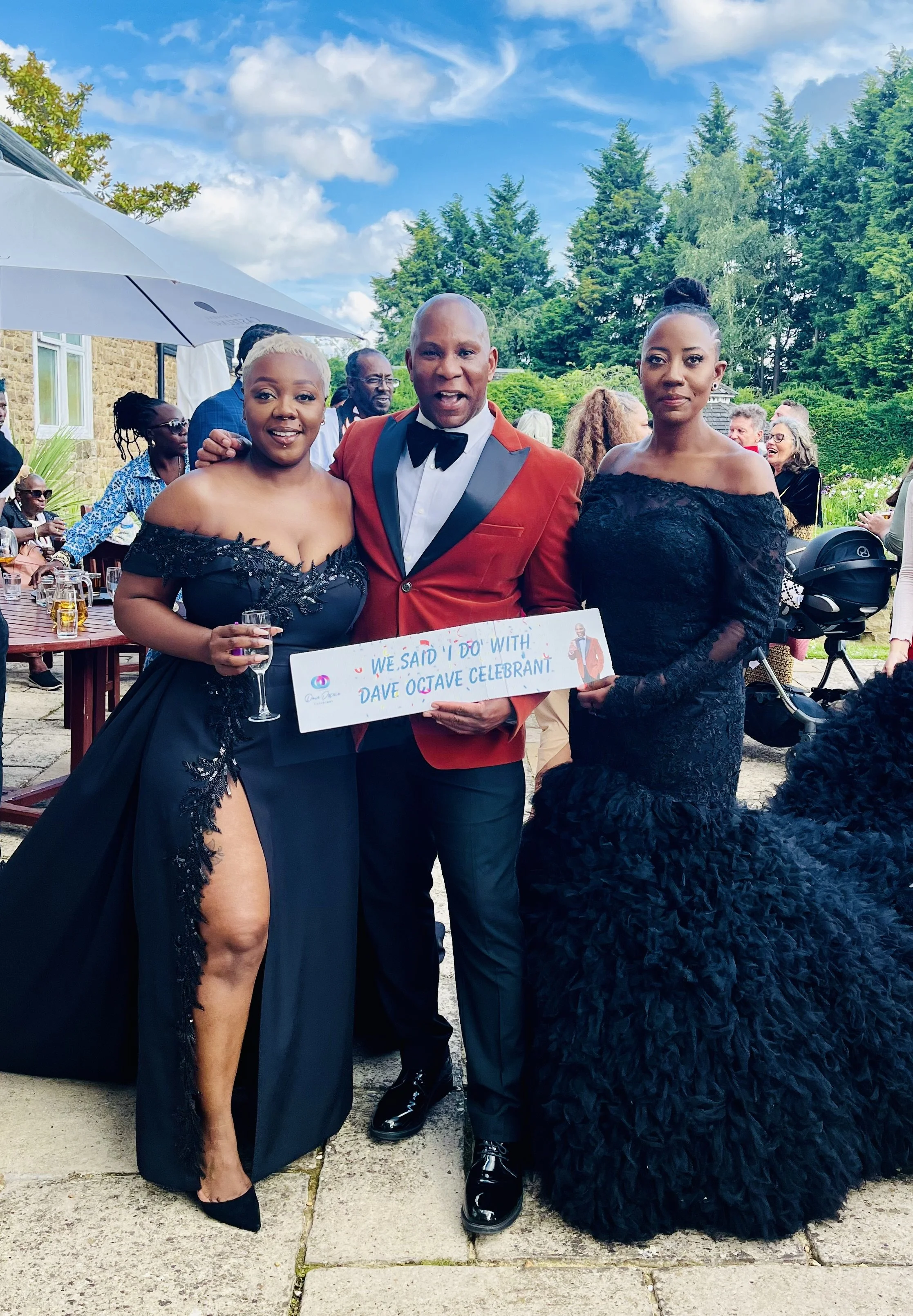 Dave Octave Celebrant in a red blazer, with 2 brides, holding a sign reading 'We said "I do" with Dave Octave Celebrant". 