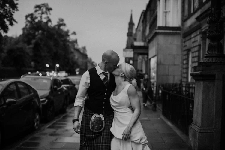 A candid black-and-white wedding portrait captured by Mirrorbox Photography, showing a couple sharing a kiss on a quiet Edinburgh street at dusk. The groom wears traditional Scottish attire, while the bride’s modern, minimalist gown adds soft contras