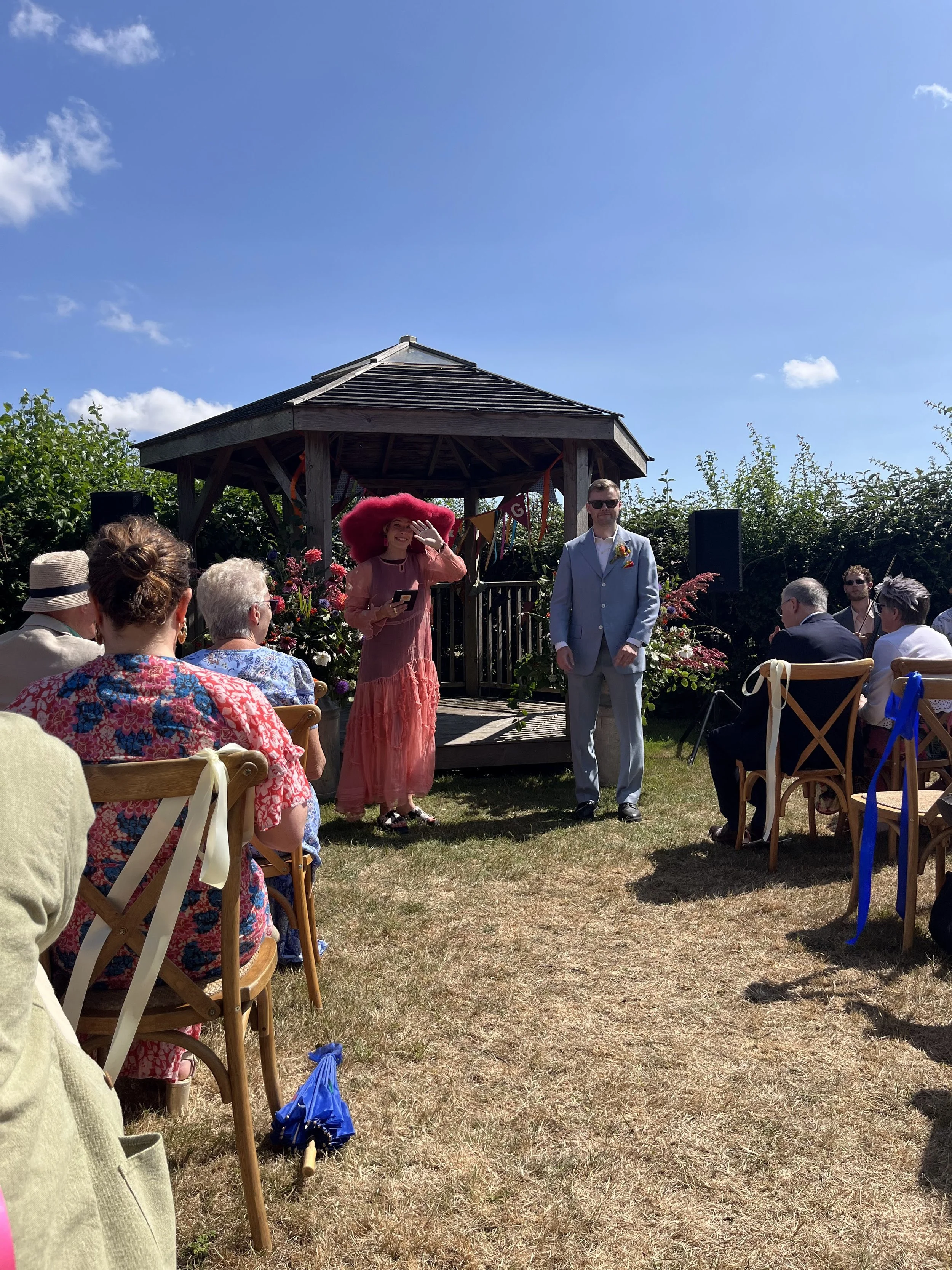 An outdoor summer wedding ceremony set under clear blue skies, led by a vibrant Tab Taylor wedding celebrant in a coral dress and bold red hat. Guests sit on wooden chairs facing a rustic gazebo decorated with bunting and colourful flowers, creating 