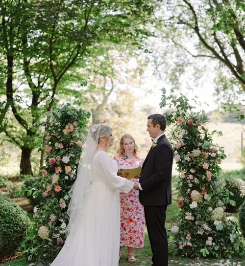 Romantic outdoor wedding ceremony set in a lush garden with the couple standing beneath floral arches. The bride wears a flowing white gown and veil, while the groom is dressed in a classic black suit. The celebrant, in a floral dress, stands between