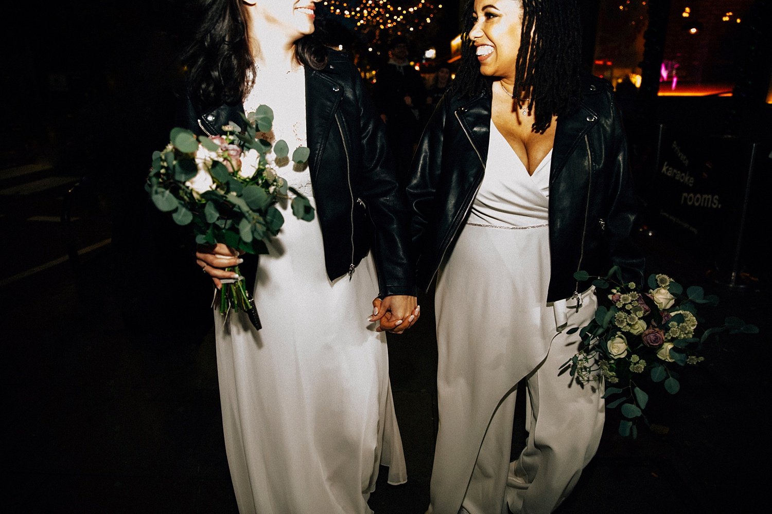 A night-time rush of joy as the two brides stride hand-in-hand through the city lights, completely wrapped up in each other. Both wear matching white dresses paired with badass black leather jackets, giving the whole moment an effortless, rock-and-ro