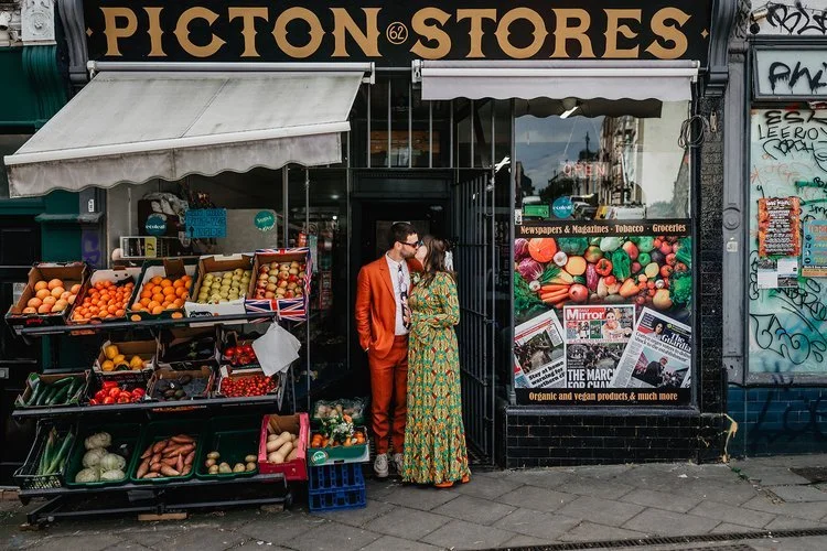 A couple share a quiet, affectionate moment outside a colourful corner shop, standing between crates of fresh fruit and bold storefront signage. Their vibrant outfits - an orange suit and a patterned green dress -blend seamlessly with the lively stre