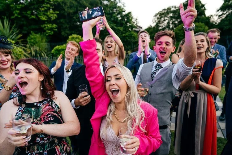 Wedding guests celebrate with huge smiles, raised glasses and lively cheers during an evening reception, captured brilliantly by Jordanna Marston Photography. The vibrant colours, expressive reactions and upbeat atmosphere create a fun and dynamic sc