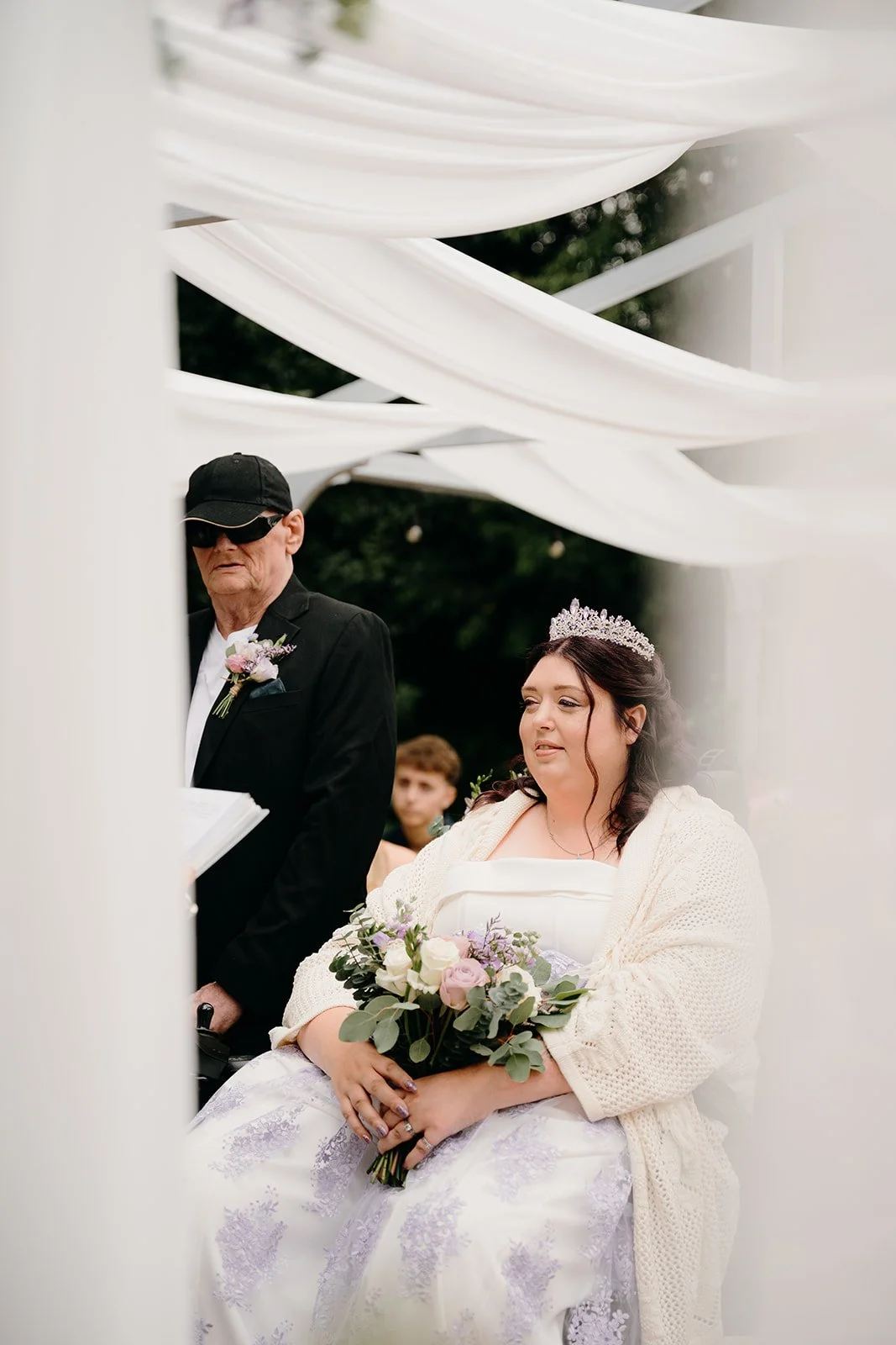 A wedding photo of a disabled bride and groom at their wedding ceremony. The romantic shot has been taken from beyond some soft, white drapes. You can see the bride in her lilac and ivory wedding outfit, holding a bouquet. The groom is at her side.