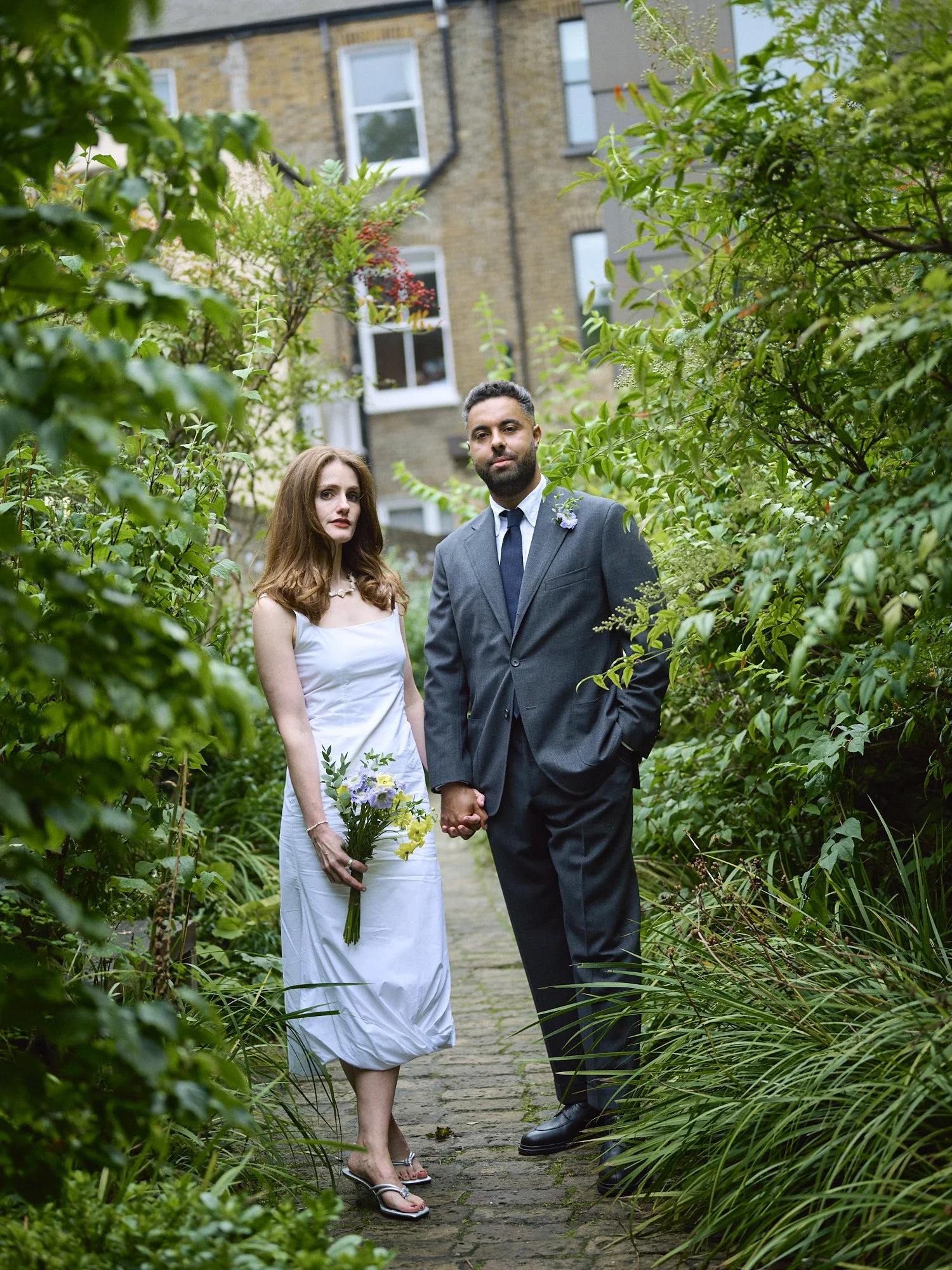 A quietly striking wedding portrait captured by Wayne La Photography, showing the couple standing hand in hand along a narrow garden pathway surrounded by lush greenery. The groom’s charcoal grey suit and composed stance contrast beautifully with the
