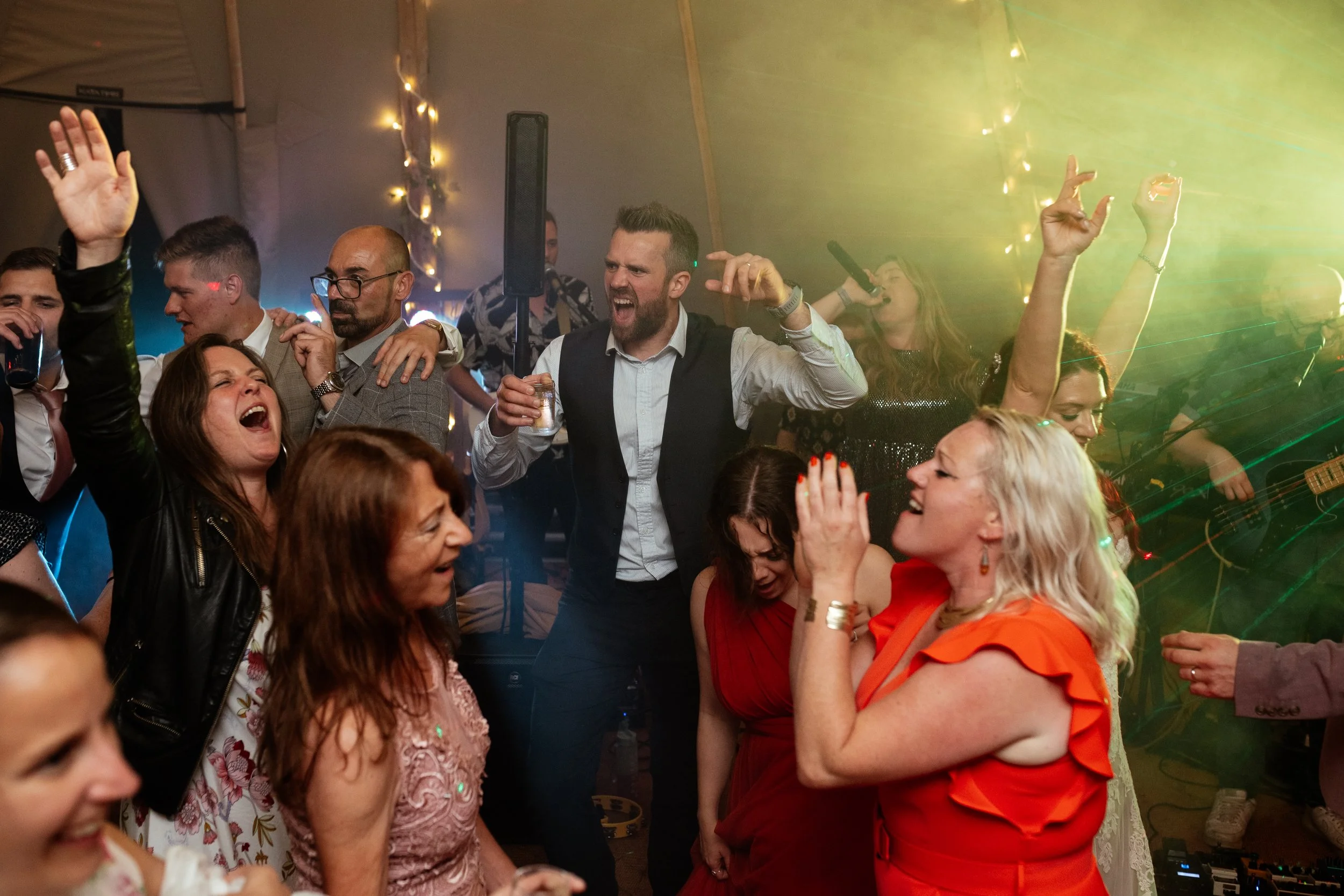 A high-energy wedding party moment captured by Laura Wilson Photography, showing guests singing, dancing and celebrating inside a tipi reception filled with colourful lights and music. People raise their hands, laugh and shout along to the band perfo