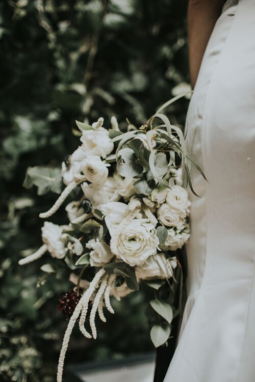 Elegant white bridal bouquet by Catherine Short Florals featuring garden roses, ranunculus, amaranthus and soft eucalyptus in a cascading natural style. Shown against a sleek bridal gown with lush greenery in the background, this refined modern arran