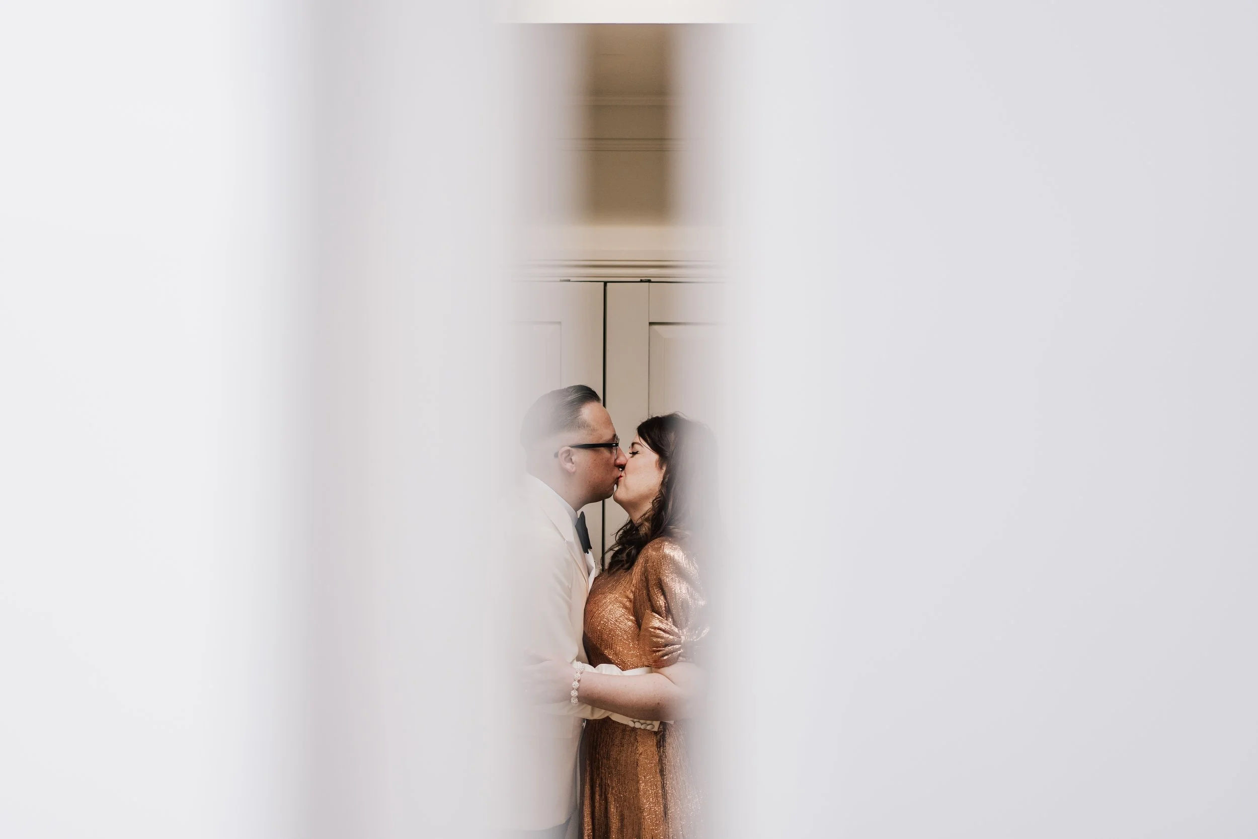 A photo of a modern bride and groom kissing. It has been captured by their wedding photographer through a crack in the door.