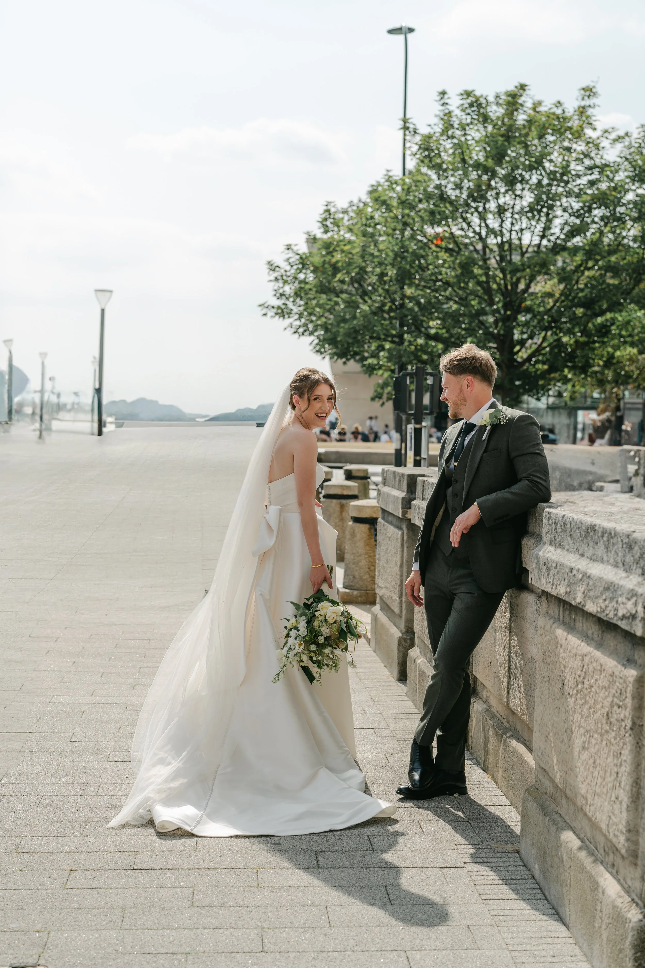 A bride and groom share a relaxed moment along a sunlit waterfront promenade, captured beautifully by Christy Photography. The bride’s elegant gown, flowing veil and soft bouquet contrast with the textured stone wall as the groom leans toward her, cr