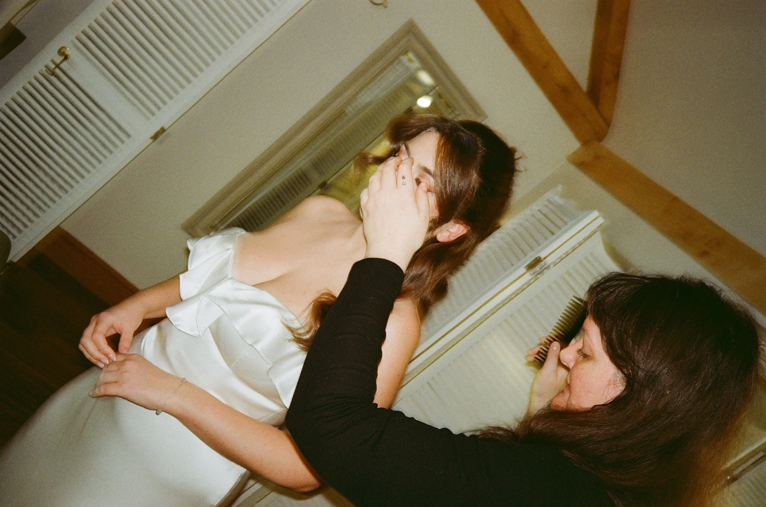 A candid wedding morning moment shows a bride in a silky white dress having the final touches done to her hair and makeup by a stylist. Photographed on 35mm film by Teddy Photo + Film, the image captures natural, documentary-style bridal prep with so