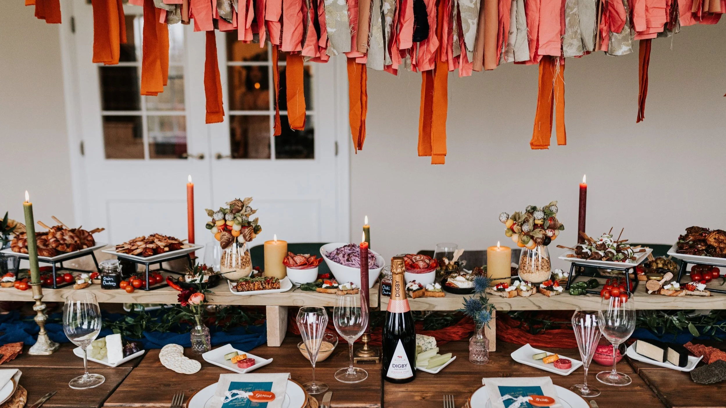 A vibrant wedding grazing table styled with bold coral and terracotta hanging ribbon decor, featuring an abundant display of artisanal meats, cheeses, fresh vegetables, breads and dips.