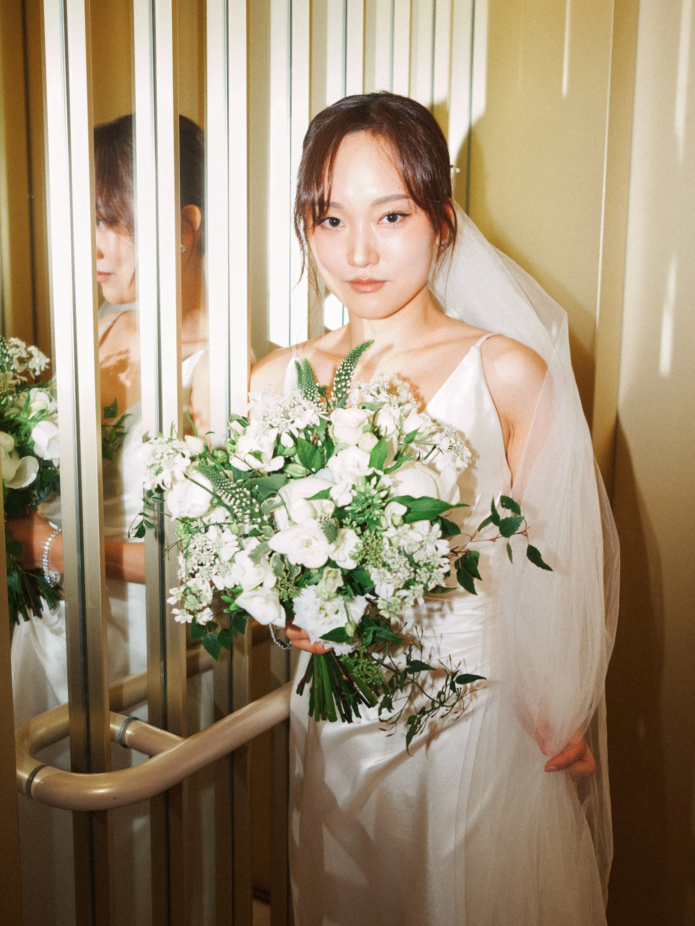 A stylish and modern wedding portrait captured by Wayne La Photography, showing the bride illuminated softly as she stands in a small elevator space, bouquet in hand. The reflective panels beside her create a subtle double image, adding a contemporar
