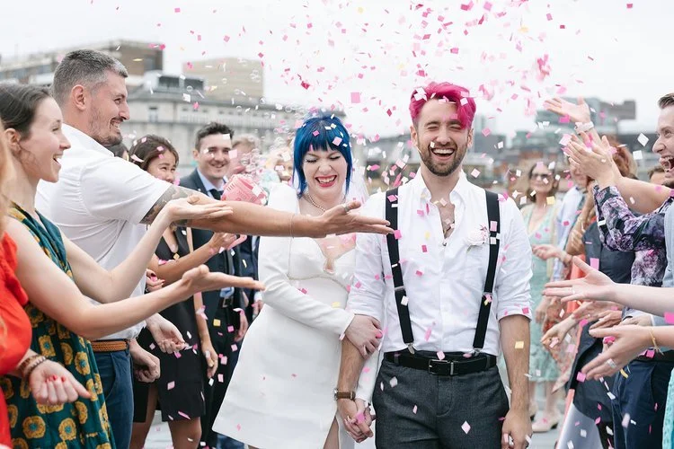 A newly married couple walk through a joyful confetti tunnel surrounded by cheering guests, captured beautifully by Christy Photography. Bright colours, laughter and movement fill the scene as the couple celebrate together, creating an energetic and 