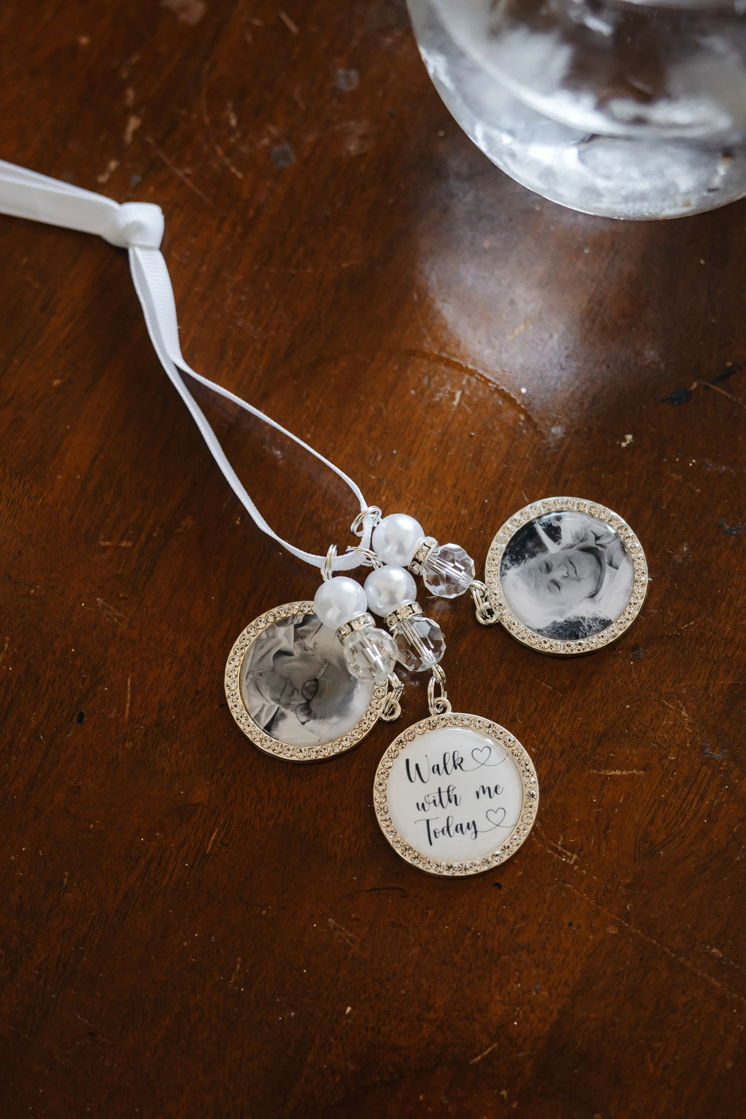 A pearl and crystal bridal bouquet charm with photo pendants rests on a wooden table. Two hold black and white portraits, and one reads “Walk with me Today,” as a sentimental aisle keepsake.