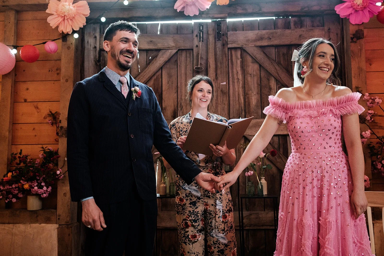 A joyful barn wedding ceremony led by Cherry and Fern Celebrations, featuring a bride in a playful pink off-the-shoulder gown and a groom in a navy suit. The rustic wooden backdrop is decorated with vibrant paper flowers, pink lanterns, and seasonal 