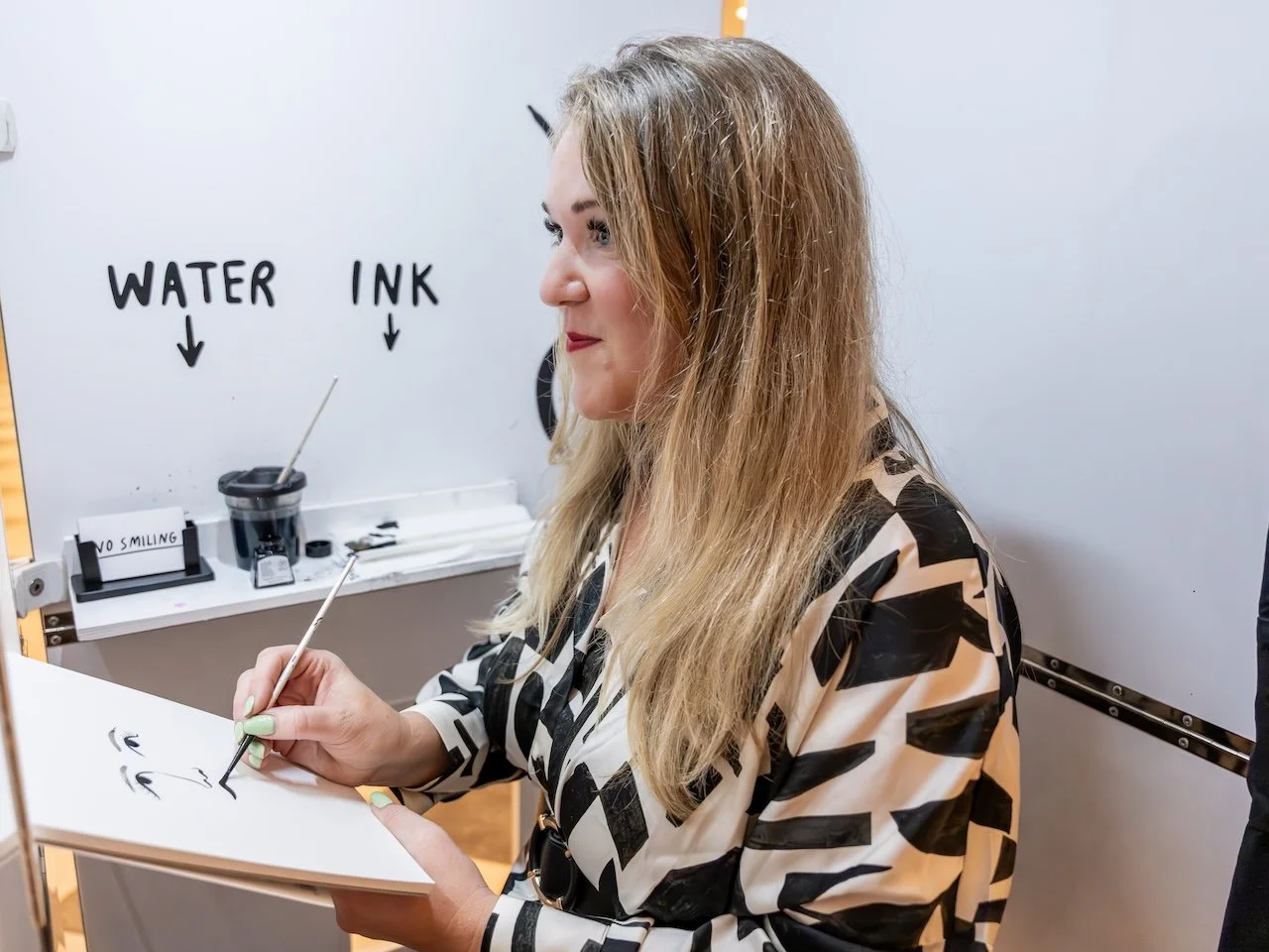 Lucie Sherican aka Rubish Portraits creating a live ink portrait inside the ‘Rubbish Portraits’ booth. Using a fine brush and minimalist black ink, she paints expressive faces on white paper with a playful ‘Water’ and ‘Ink’ sign behind her. Captured 