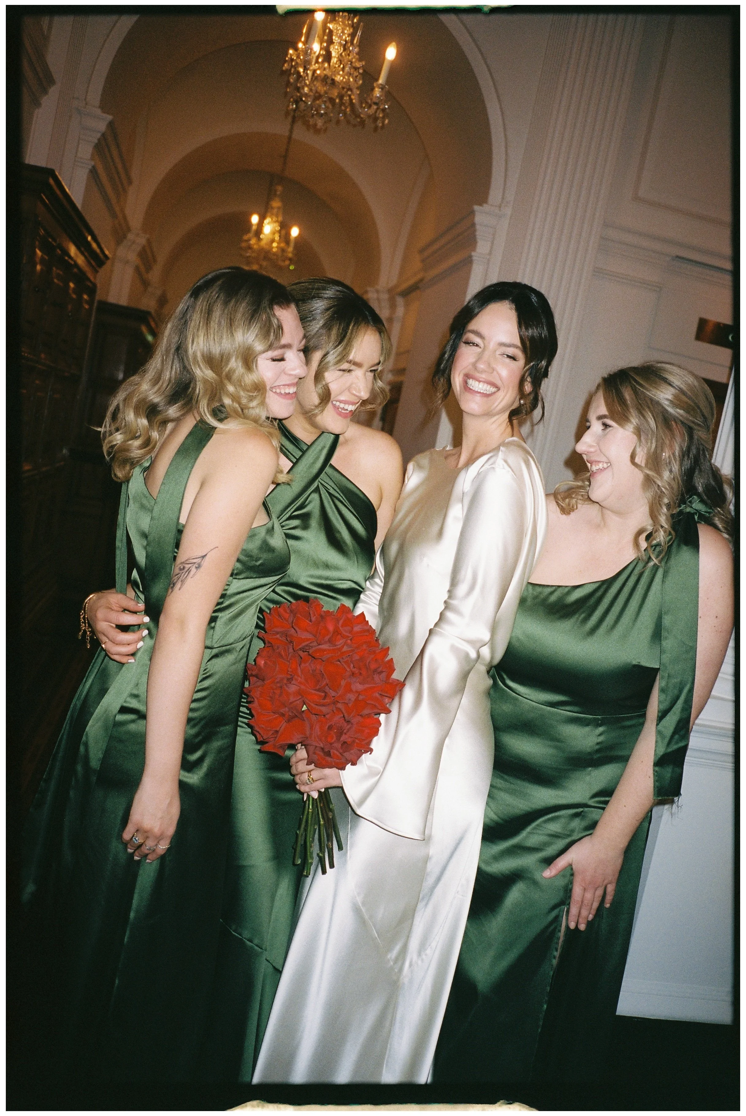 A radiant bridal party captured in timeless elegance, featuring the bride in a lustrous ivory satin gown surrounded by bridesmaids wearing emerald green satin dresses by Rewritten Bridal Wear. The dresses, each with a slightly varied neckline and dra