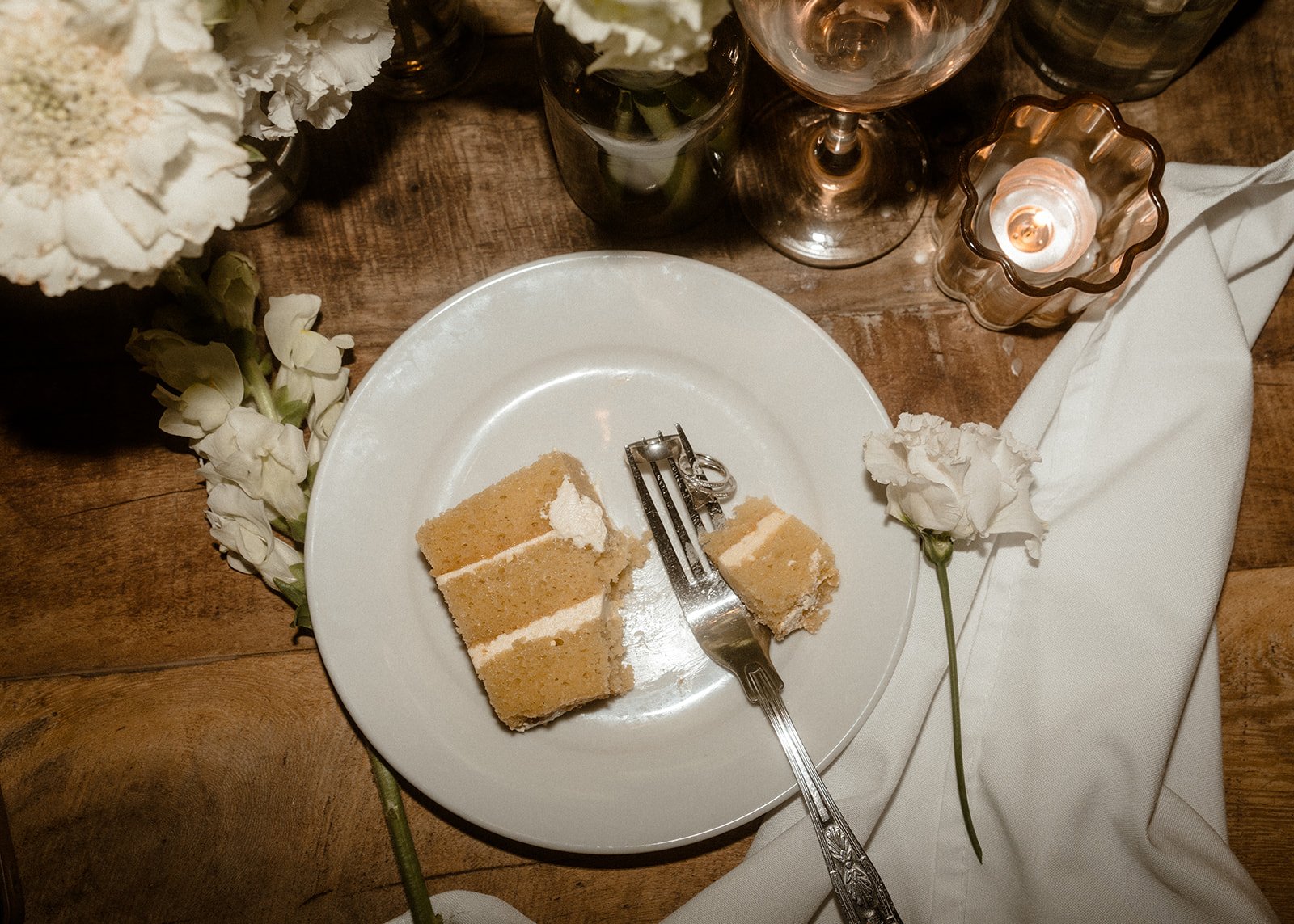 A plate with partially eaten vanilla wedding cake sits on a rustic wooden reception table alongside white flowers, a lit candle, a wine glass and a folded white napkin with a silver fork resting on the plate.
