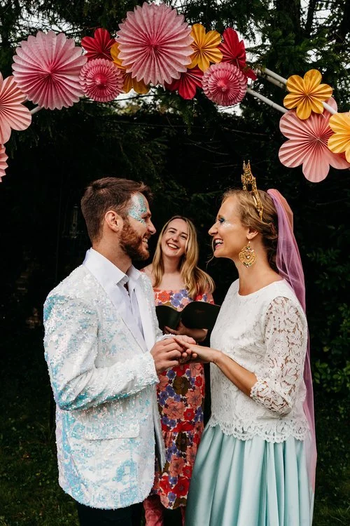 A fun and colourful outdoor wedding ceremony led by smiling UK celebrant Frances Celebrates You. The couple stand beneath a bright arch of paper flowers, exchanging rings and happy glances. The bride wears a pastel skirt, lace top and pink veil with 