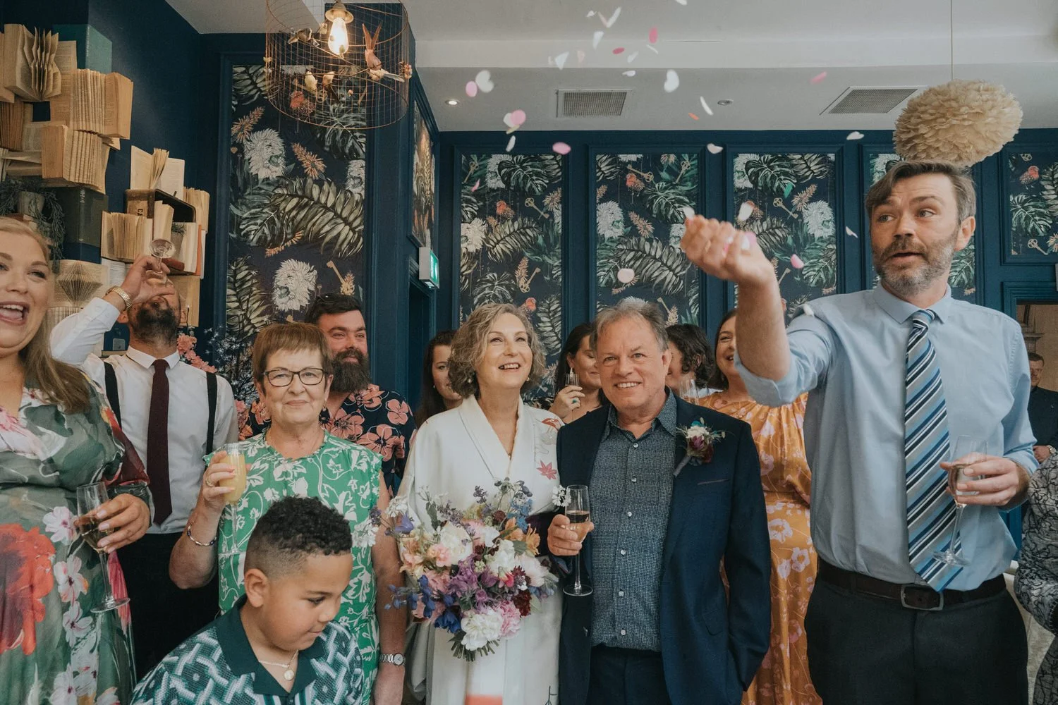 A group of wedding guests gathers indoors beneath patterned wallpaper as confetti floats through the air. At the center, an older couple smiles warmly, holding drinks, while others around them laugh, cheer, and raise glasses in celebration. The scene