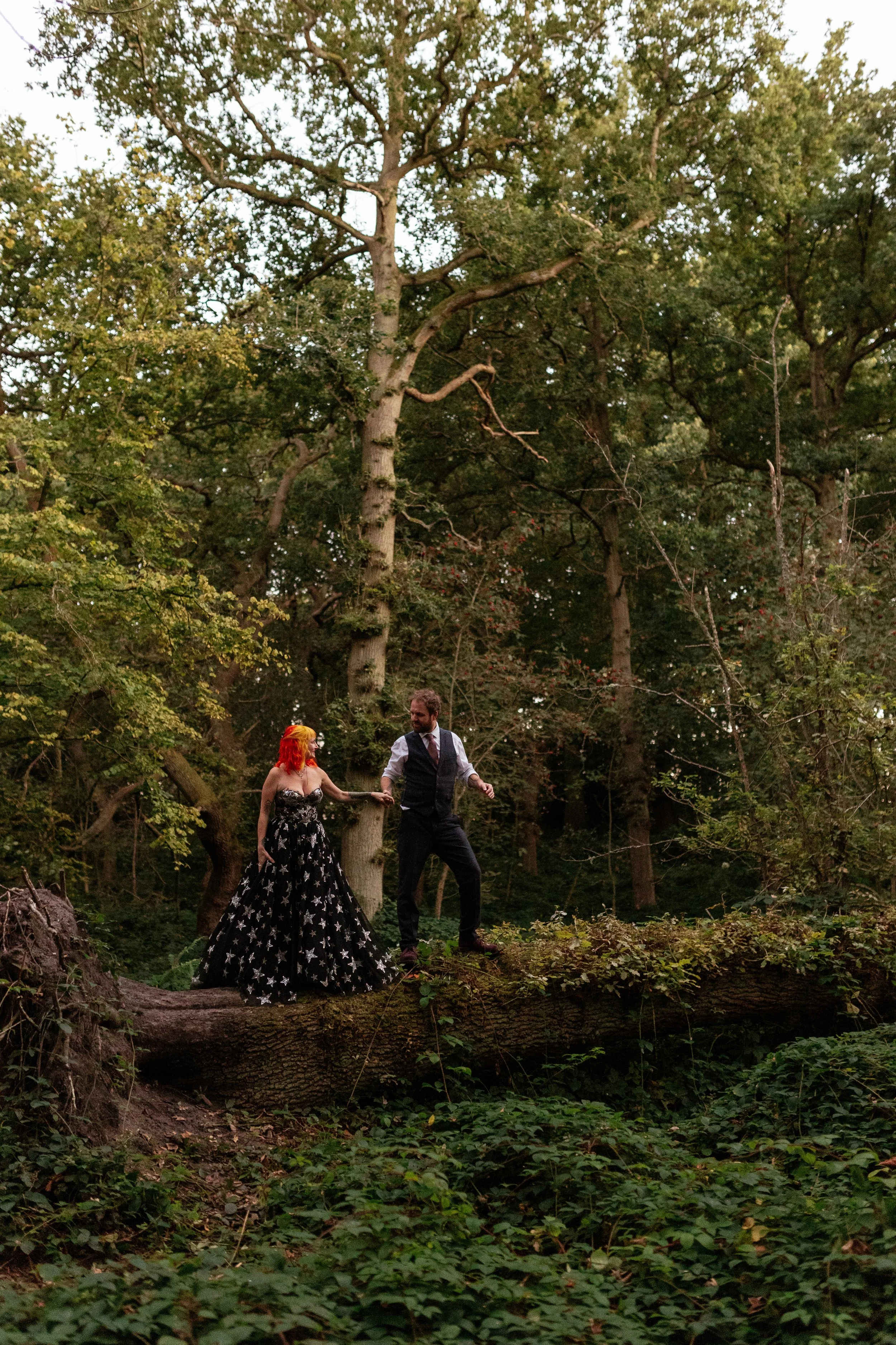 A creative and dramatic wedding portrait captured by Laura Wilson Photography, showing a couple walking together across a fallen tree trunk in the middle of a dense woodland. The bride wears a striking black gown covered in star patterns, paired with