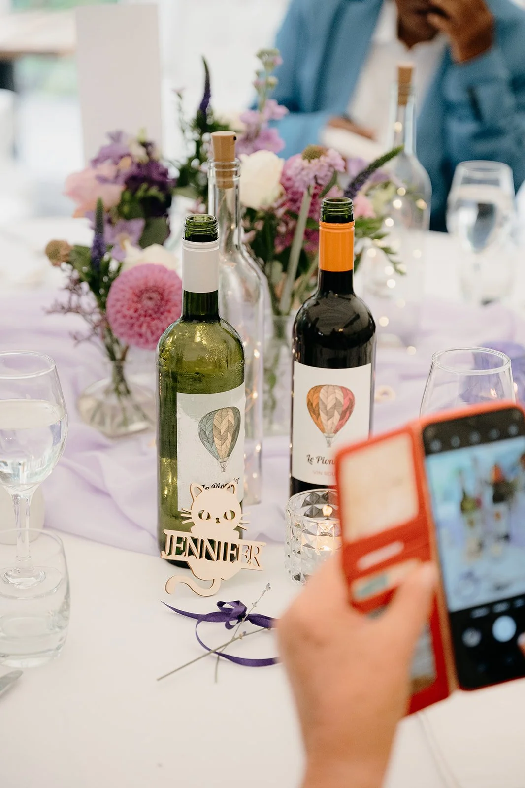 A colourful table at a wedding reception. There are flowers and bottles of wine. Someone is taking a mobile phone picture of their place setting which is a wooden cat that says 'Jennifer'.