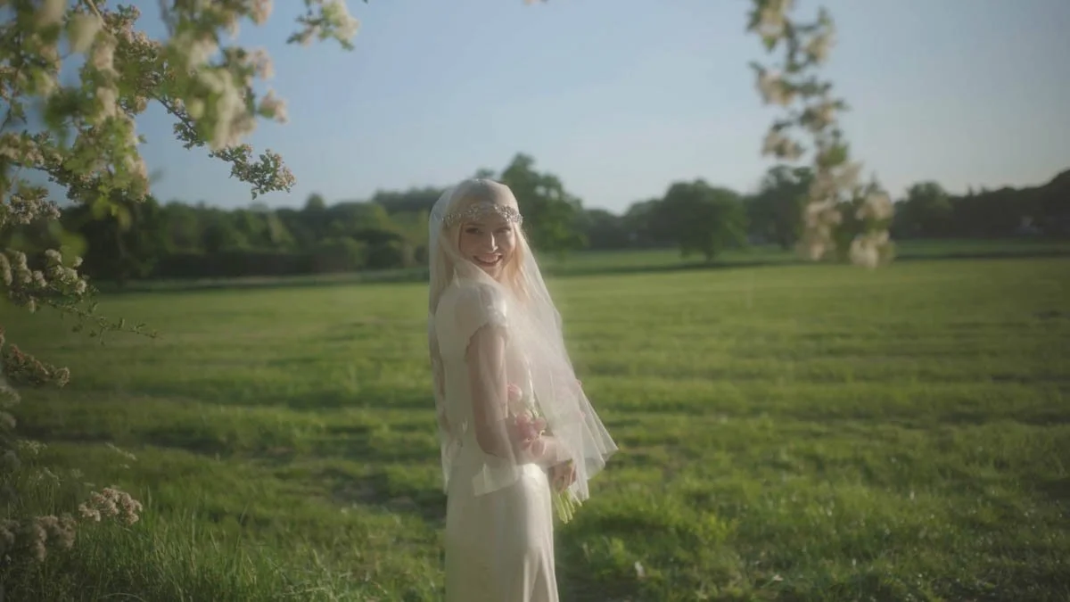 A bride stands in a sunlit meadow surrounded by soft blossoms and glowing green fields while warm evening light wraps around her delicate veil and vintage inspired headpiece as she turns back with a bright joyful smile that feels gentle cinematic and