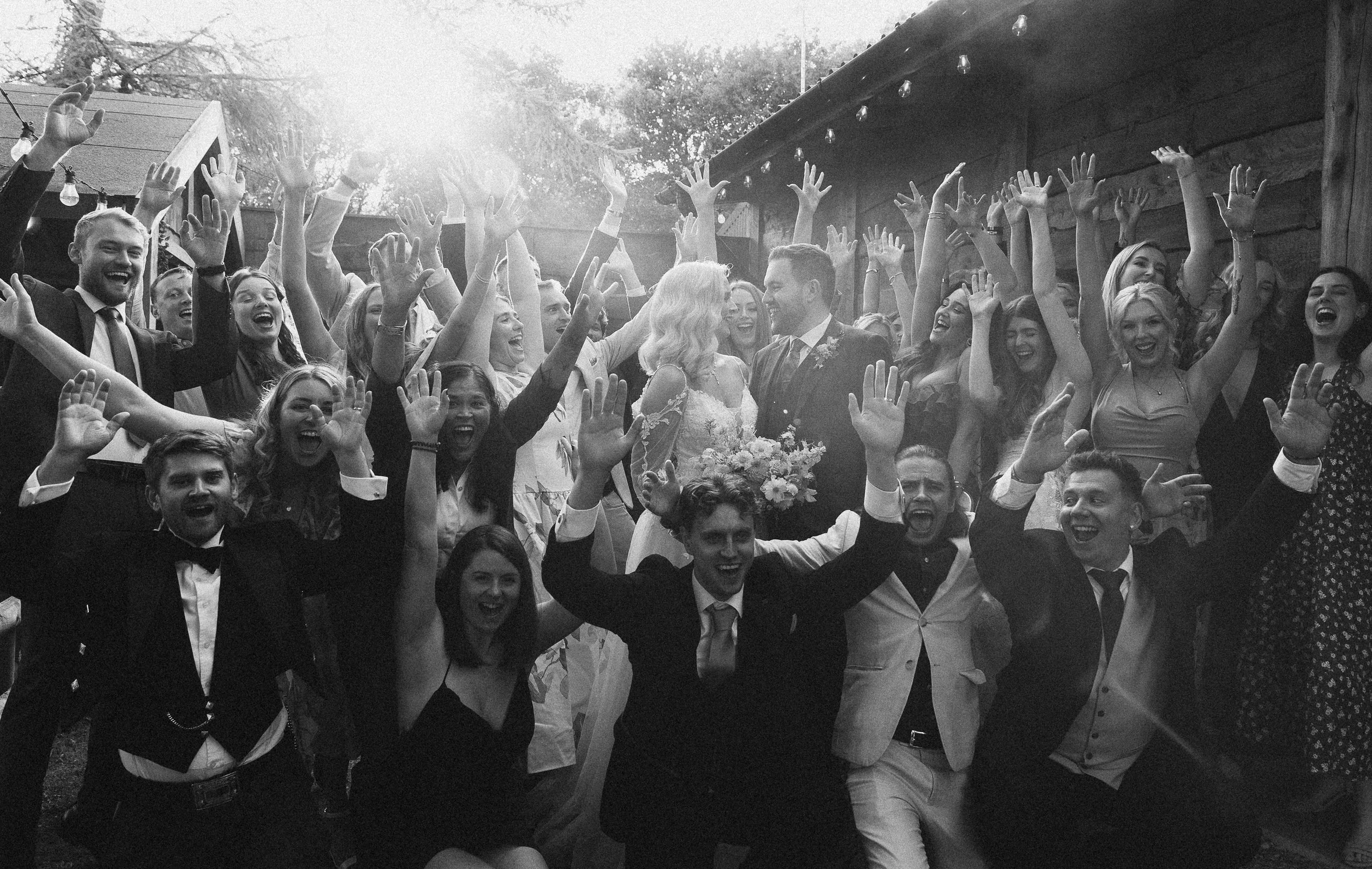 A lively and joy filled group celebration captured by YU Photography, showing the newlyweds surrounded by a sea of cheering guests with their hands thrown high into the air. The couple stand at the centre, gazing at one another with happiness as sunl