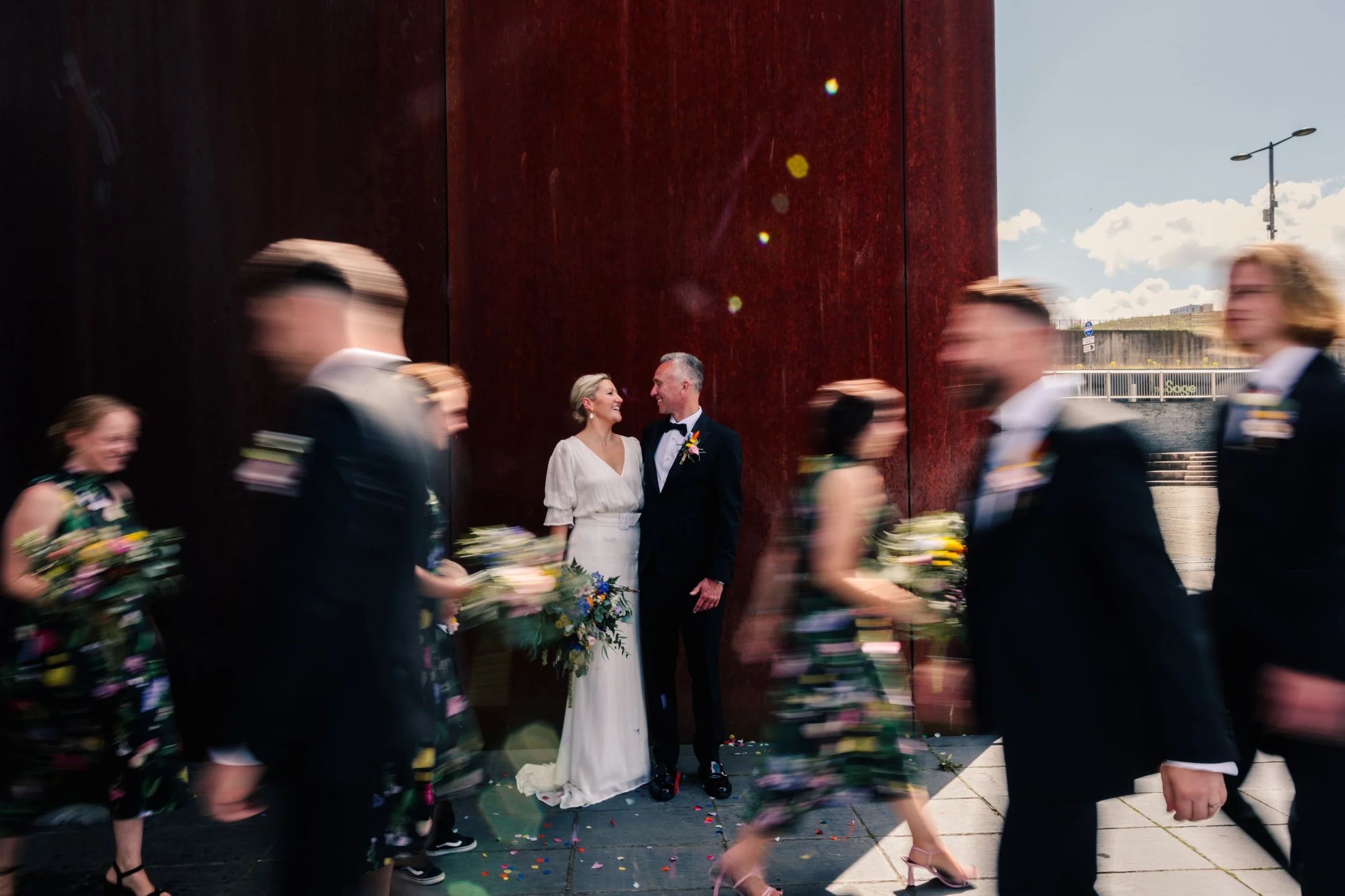A creative wedding portrait captured by Teddy Photo + Film shows a couple standing close together against a large rust-red industrial wall outside the Baltic Centre for Contemporary Art in Newcastle. The newlyweds smile at each other while their wedd
