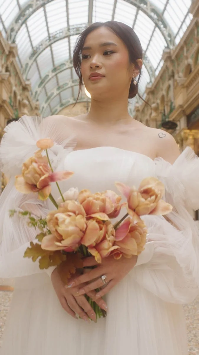 A serene bride stands beneath an ornate glass arcade holding warm peach toned blooms that echo the softness of her ethereal tulle sleeves as gentle light filters through the arched ceiling creating a graceful cinematic portrait full of quiet beauty.