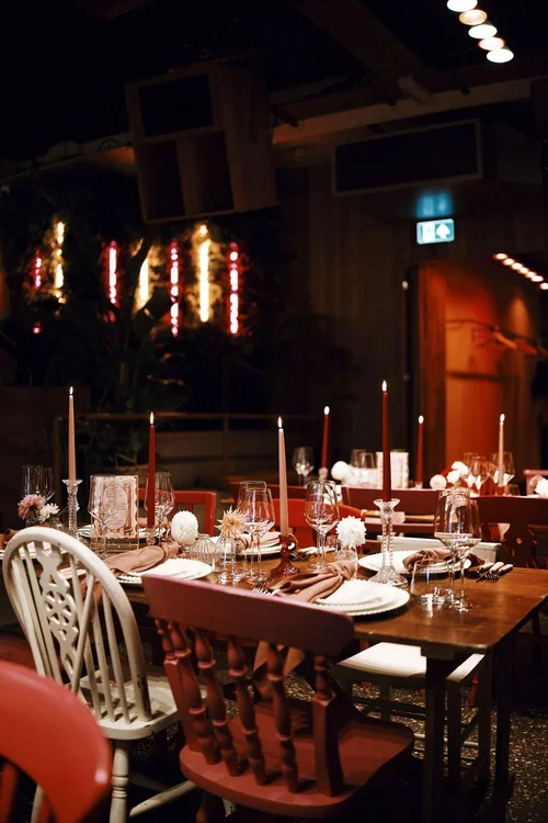 A beautifully styled wedding tablescape inside Pier Court Brighton, featuring mismatched vintage chairs in deep red tones, elegant taper candles, and modern glassware. The ambient lighting and industrial-chic interior create a warm, romantic atmosphe