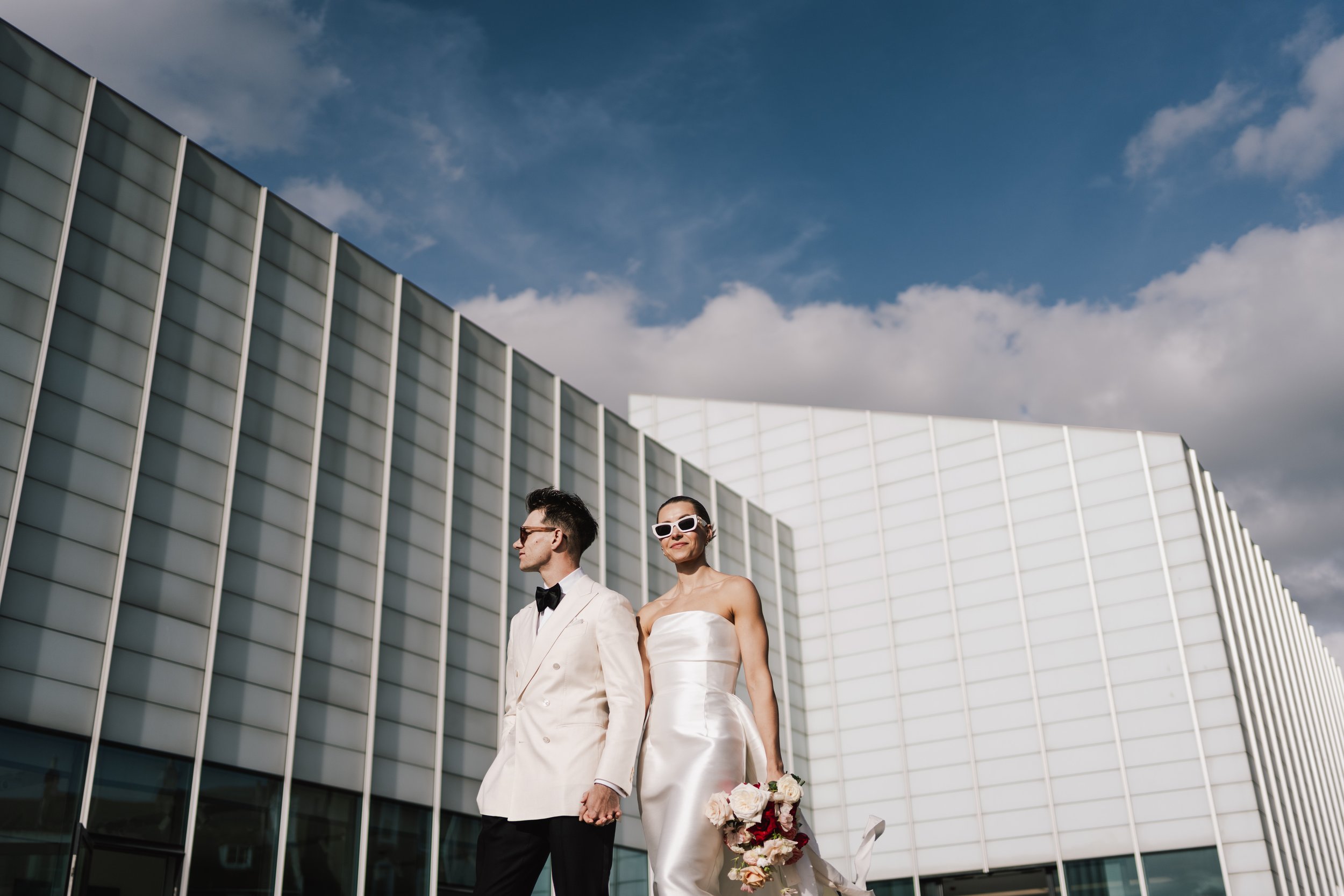 A stylish bride and groom are holding hands and walking past the Tate Modern. There is a blue cloudy sky in the background. 