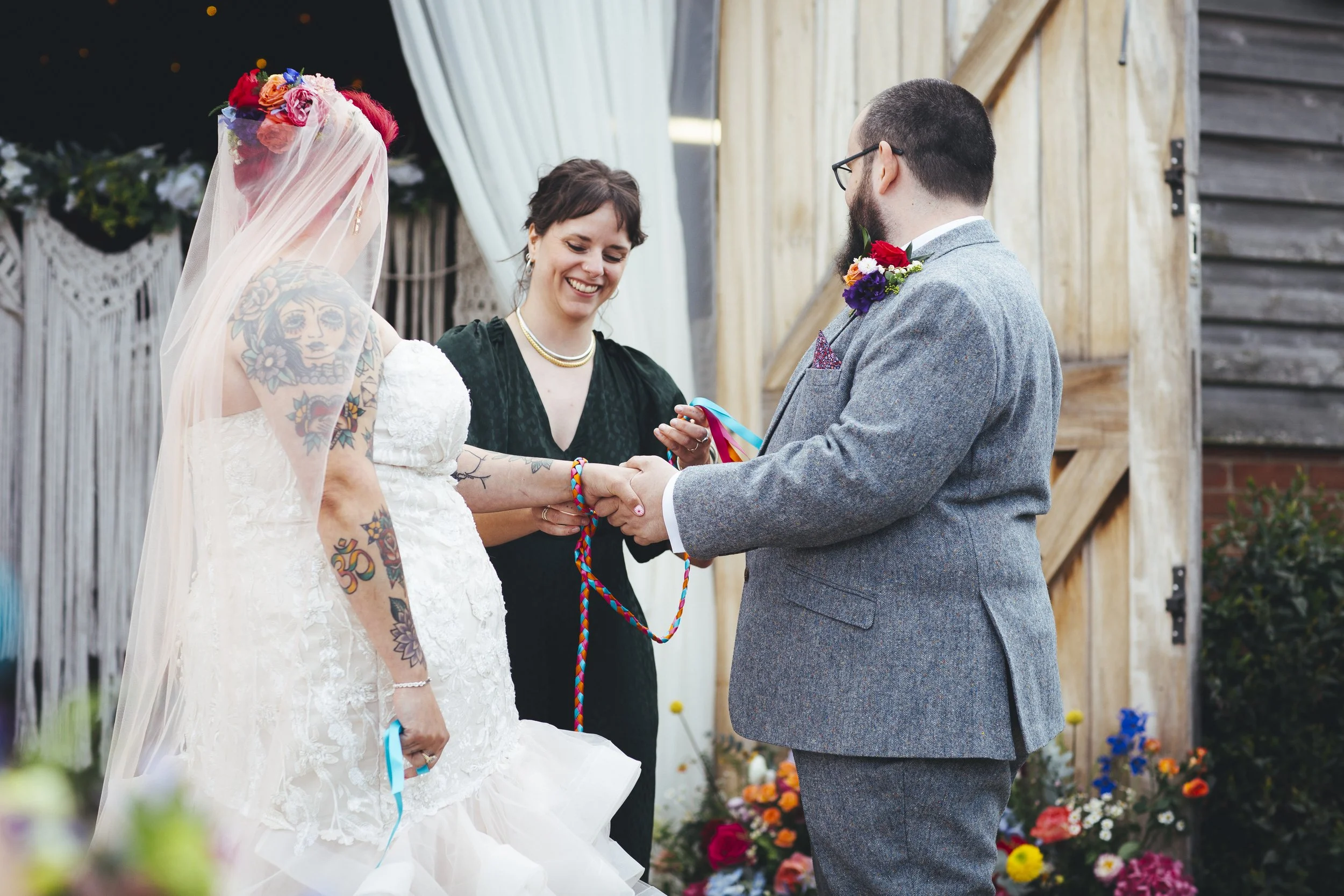 A joyful outdoor wedding ceremony featuring a tattooed bride in a lace gown and a groom in a grey suit, joined by a smiling Cherry and Fern Celebrations performing a colourful hand-fasting ritual. The rustic barn backdrop, bright floral arrangements,