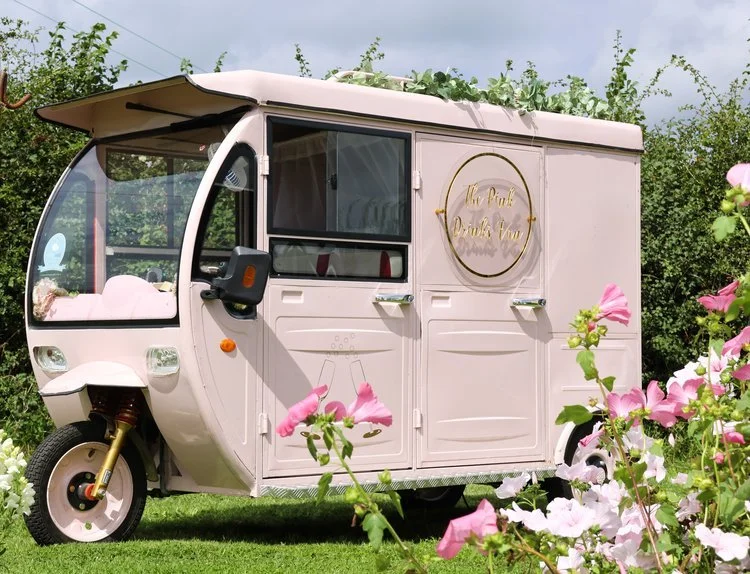 The Pink Drinks Van parked on lush green grass, surrounded by pink flowers and greenery. This pastel pink mobile bar features elegant gold signage and a stylish compact design, perfect for weddings, garden parties, and outdoor celebrations. The setup