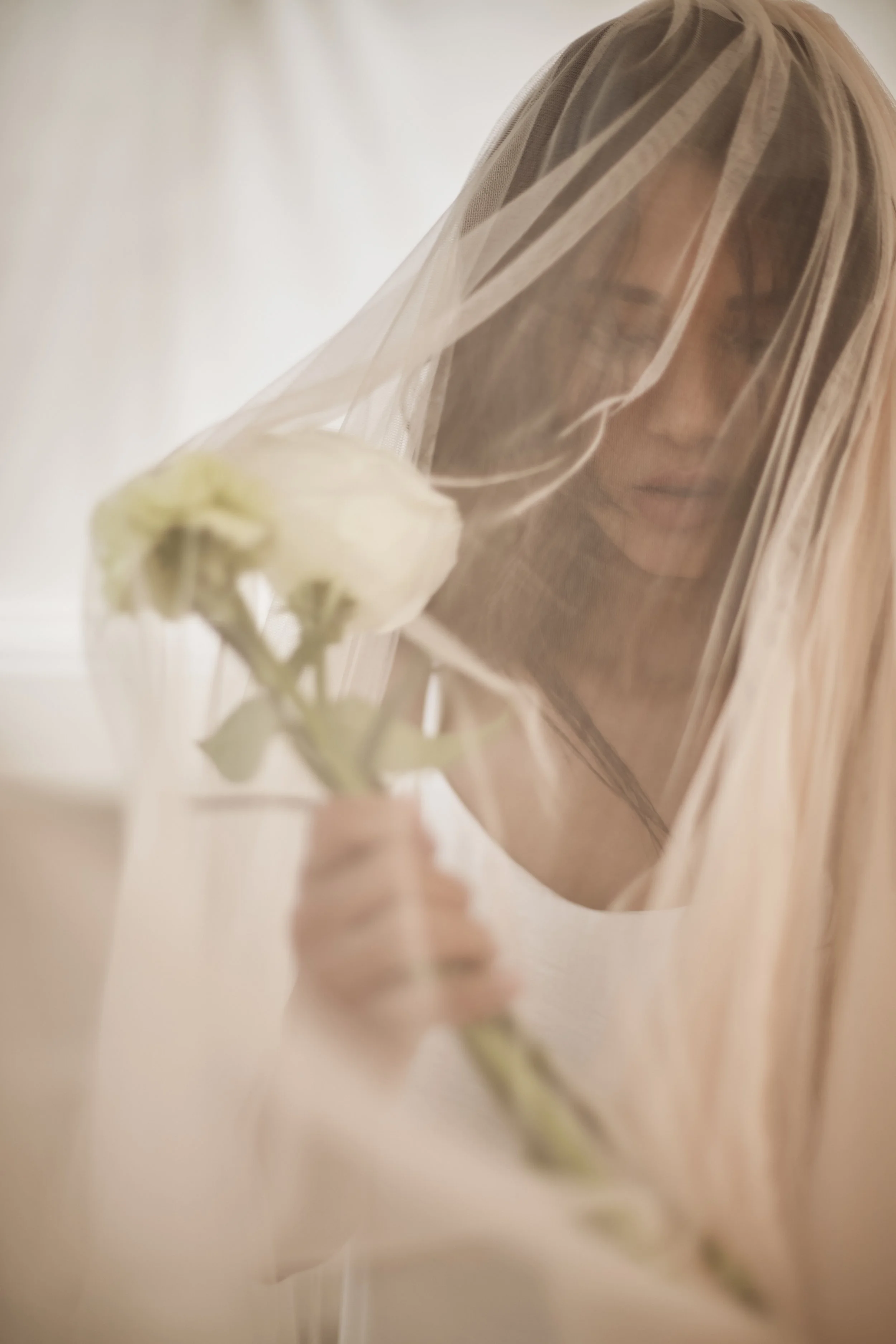 A close-up shows a person wearing a light, sheer veil that softly obscures their face as they hold a single pale flower near their chest. The image is softly focused with warm, diffused light, highlighting the translucent layers of fabric and the gen