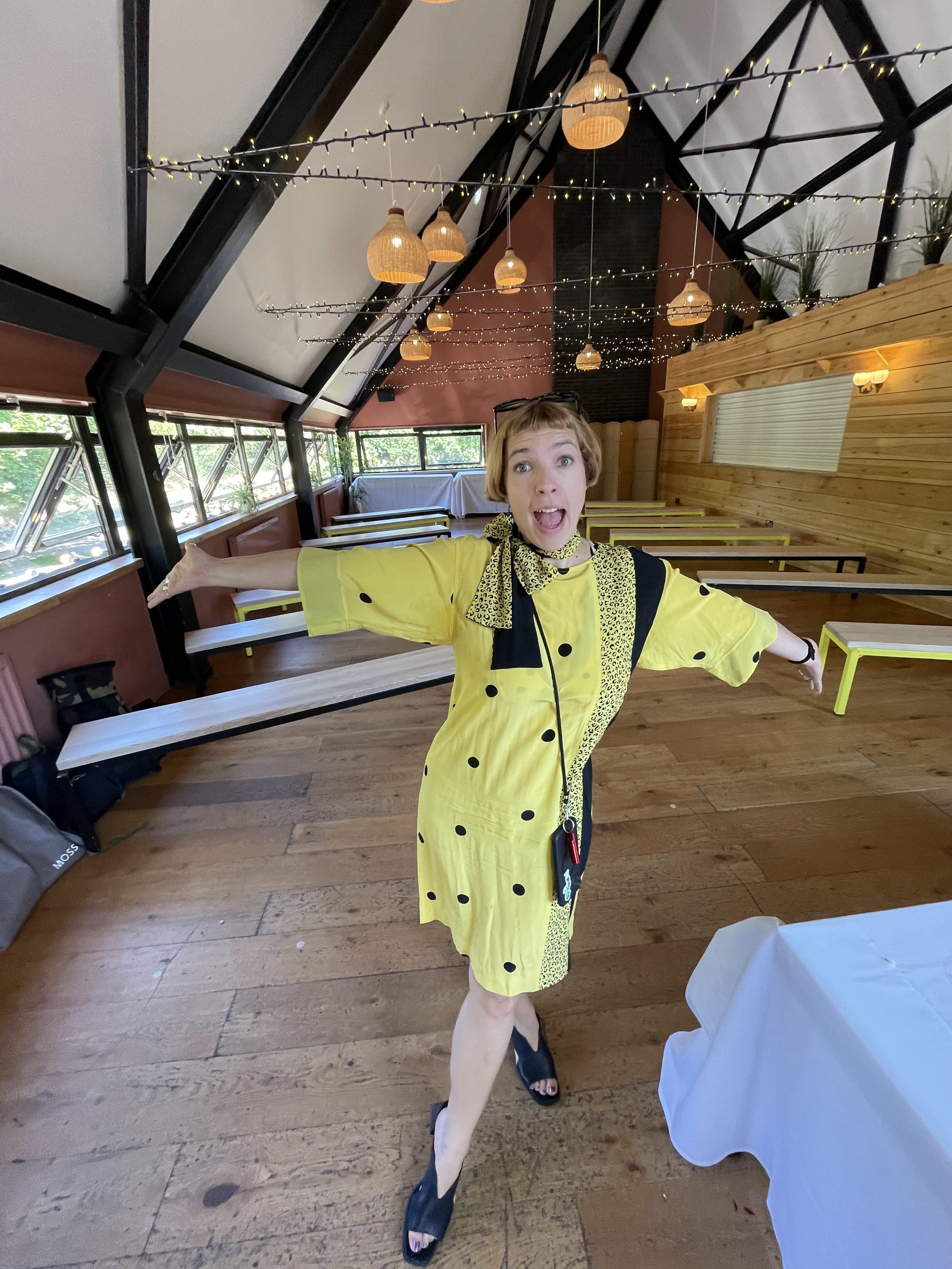 A fun, candid photo of Tab Taylor, wedding celebrant from Say it Right Ceremonies, in a bright yellow polka-dot dress and scarf, posing playfully inside a rustic modern wedding venue. The space features wooden floors, fairy lights, hanging lamps and 