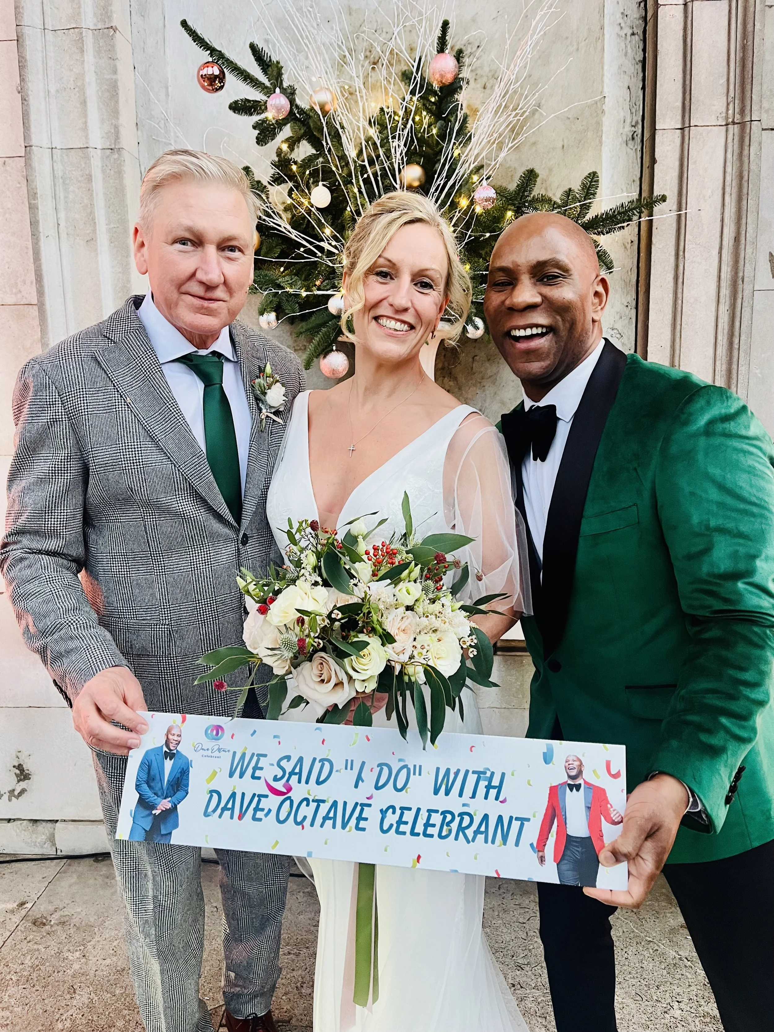 Dave Octave Celebrant in deep green blazer, with a bride and groom, holding a sign that reads 'We said "I do" with Dave Octave Celebrant". 
