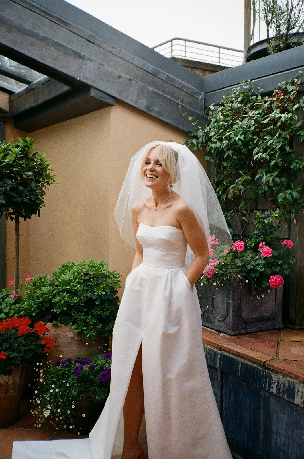 A bride in a strapless white gown with a thigh high slit and veil stands smiling in a courtyard garden area surrounded by potted plants and vibrant flowers with natural light highlighting her face.
