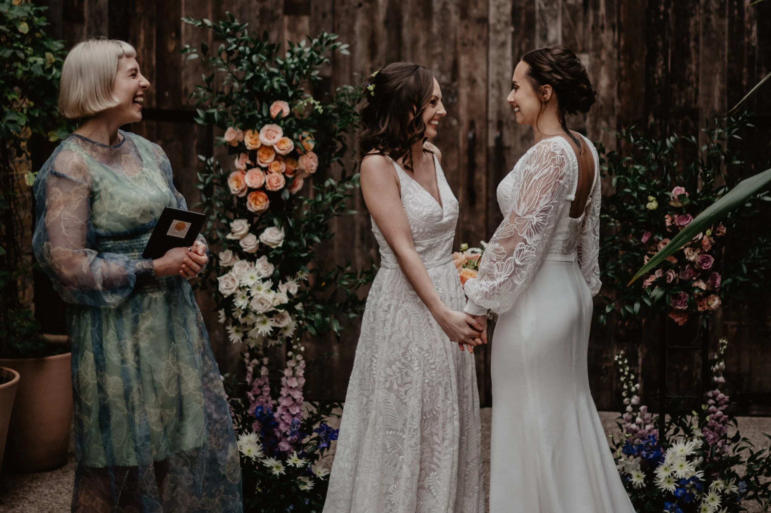 A tender moment as two brides hold hands and look at each other lovingly. Celebrant Tab Taylor looks on, smiling in a green dress.