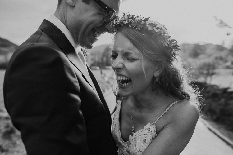 A beautifully candid wedding portrait captured by Mirrorbox Photography, showing a couple doubled over in laughter during a joyful moment together. The bride, wearing a delicate lace dress and a natural greenery crown, beams with genuine emotion as s