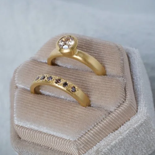 A handmade yellow gold wedding and engagement ring set by Natalie Vernazza, featuring a champagne diamond solitaire paired with a matching diamond wedding band, displayed on a soft velvet ring box.